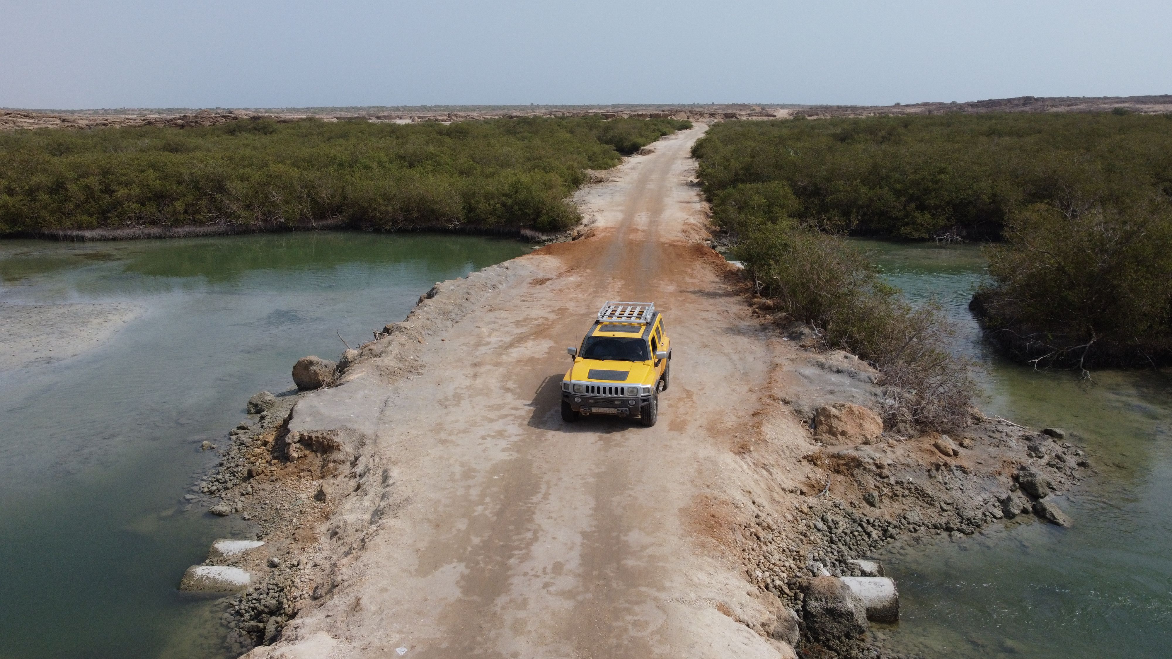 Aerial view of a yellow SUV navigating a rugged sandy road surrounded by green waterways.