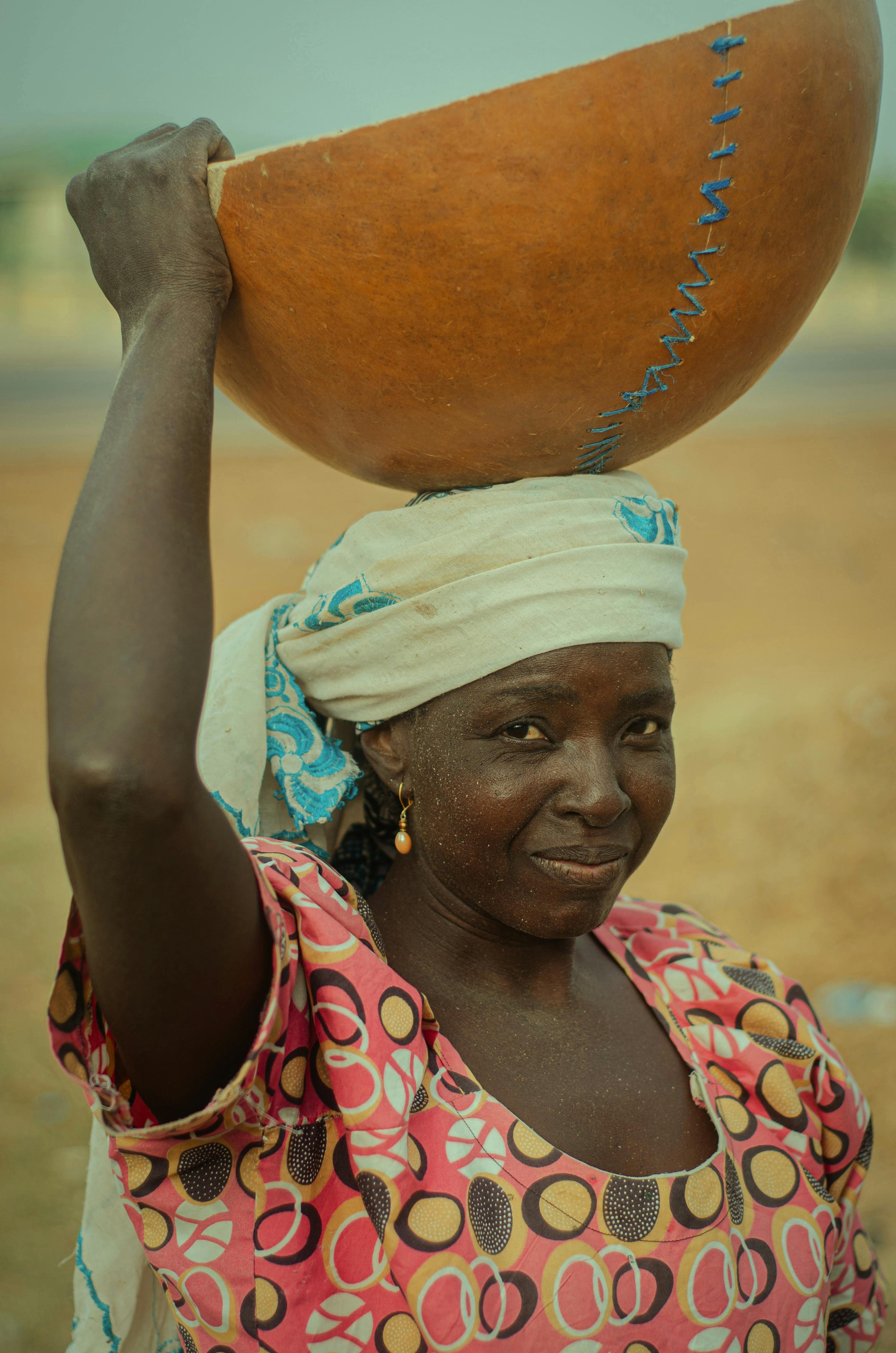 African woman with a bowl on her head, wearing patterned clothing and a headscarf in a rural setting.