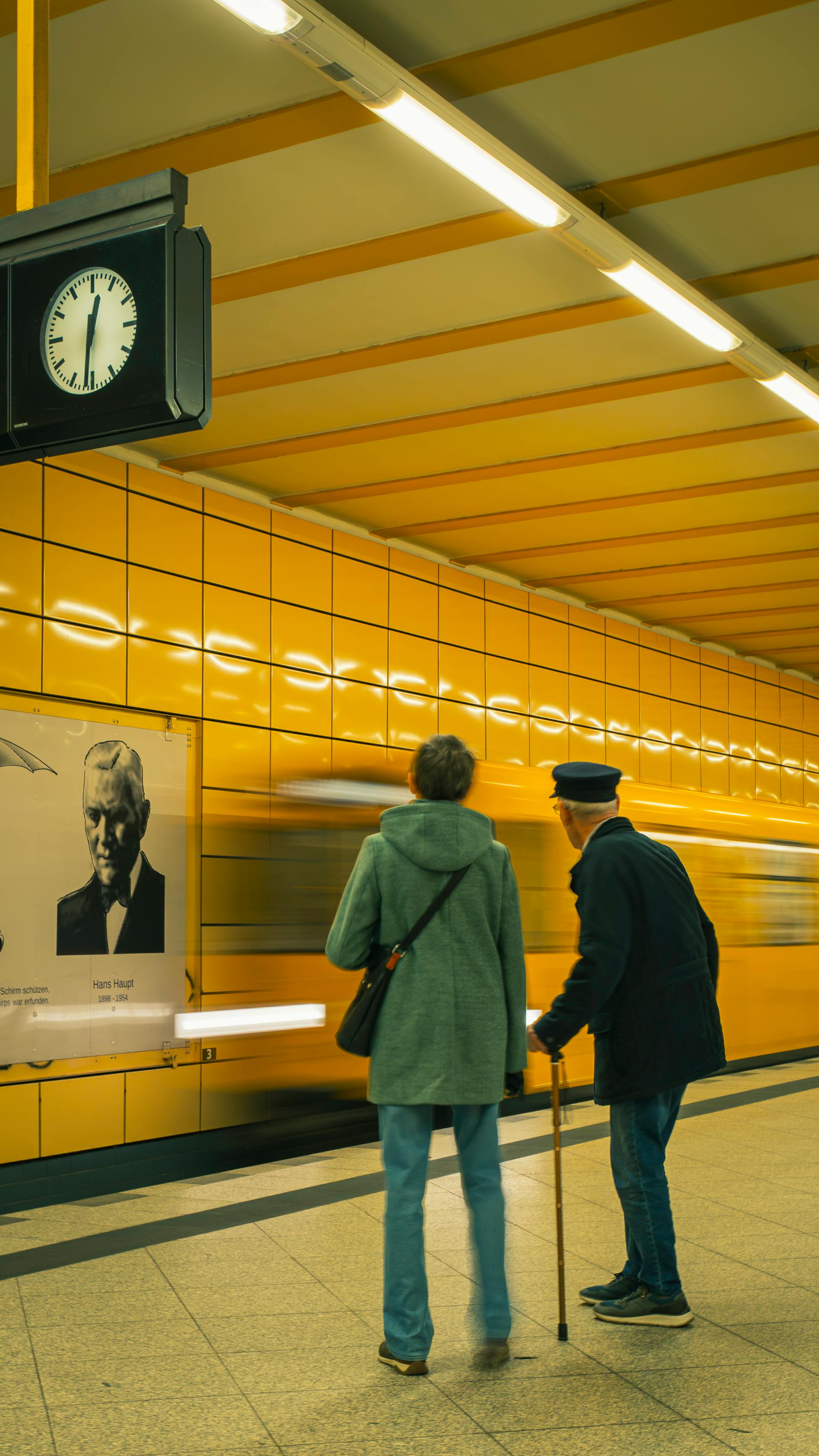 An elderly couple waits at a Berlin subway station as a train arrives. Vibrant yellow decor.