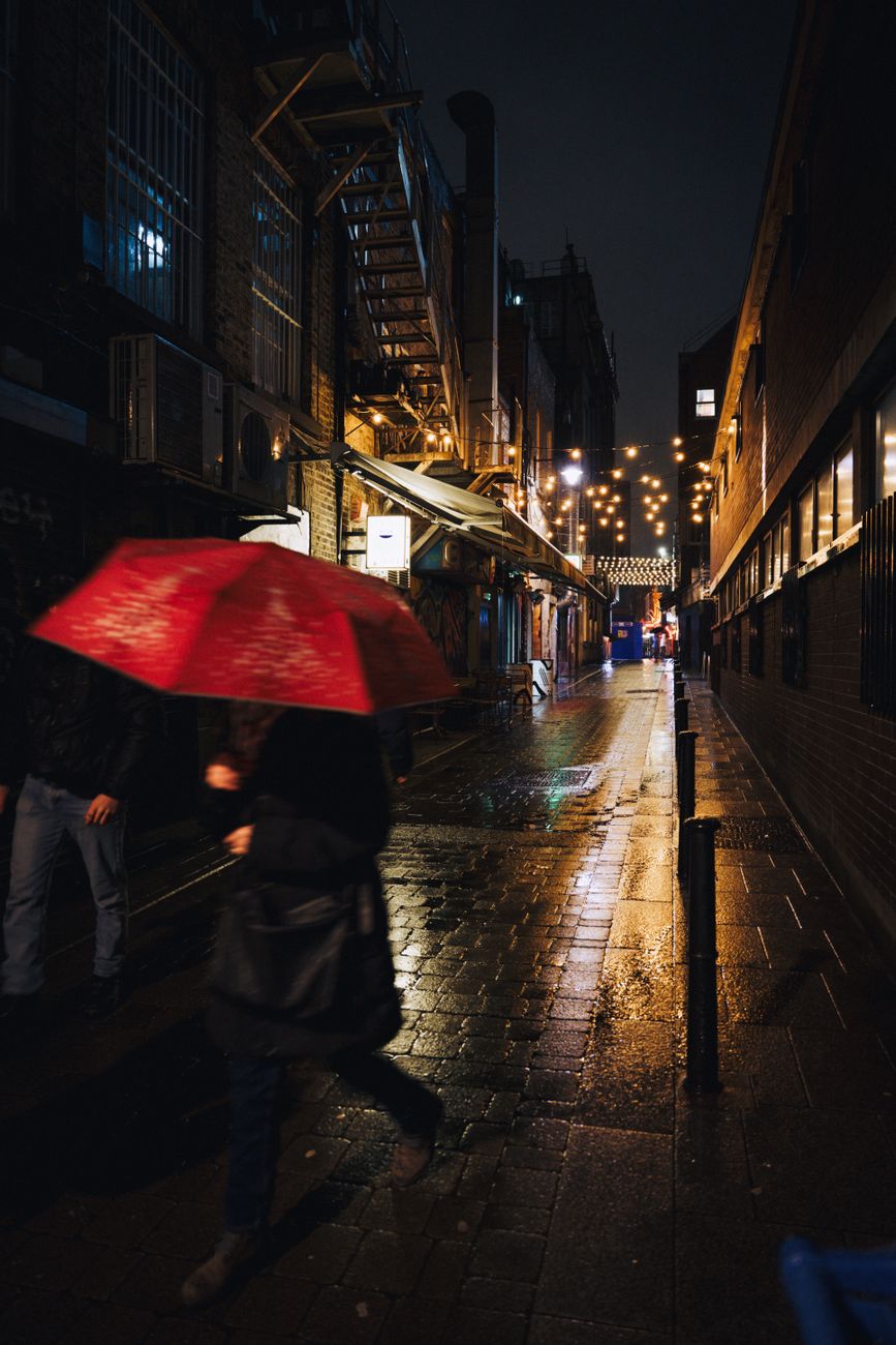 Moody street view at night featuring a red umbrella in a rain-soaked alley.