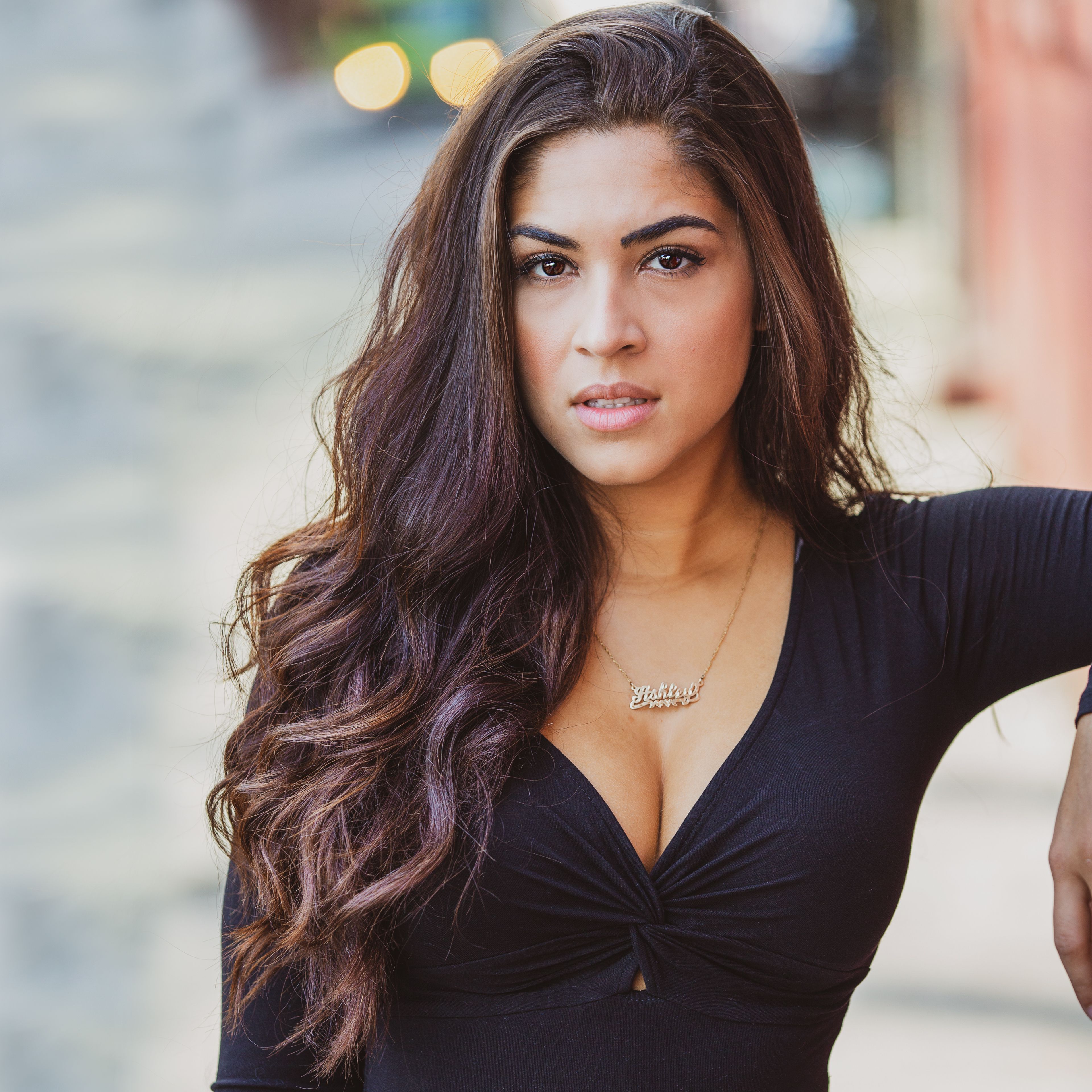 A confident young woman with long hair poses outdoors in an urban setting, exuding style and elegance.