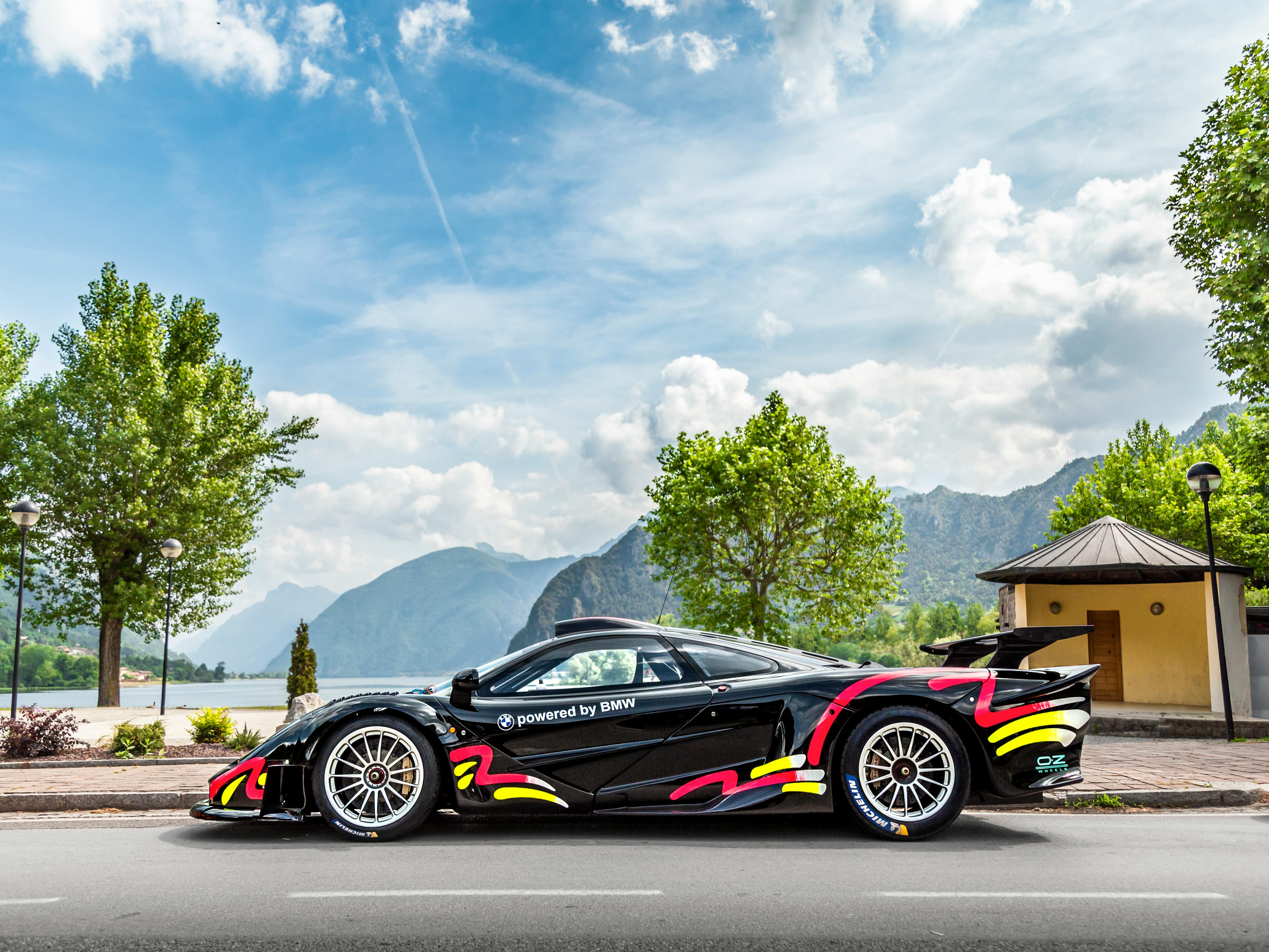 A stylish black supercar with vibrant decals parked by a picturesque lakefront with mountains in the background.