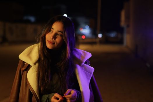 A young woman in a coat smiles softly on a dimly lit street at night.