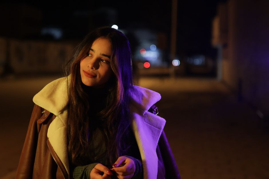 A young woman in a coat smiles softly on a dimly lit street at night.