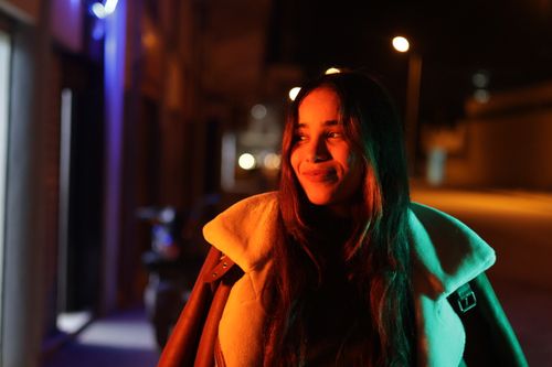 Free Smiling woman in a coat captured on a city street at night with vibrant lighting. Stock Photo