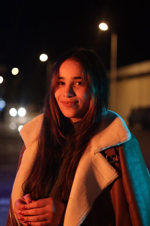 Free Young woman with long hair smiles at night, city lights softly glowing in the background. Stock Photo