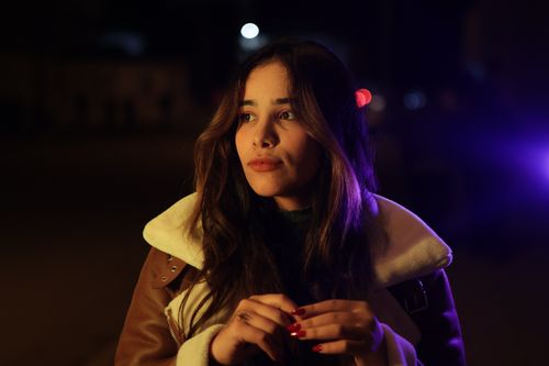 Free Woman in a brown coat with thoughtful expression under night lights. Stock Photo