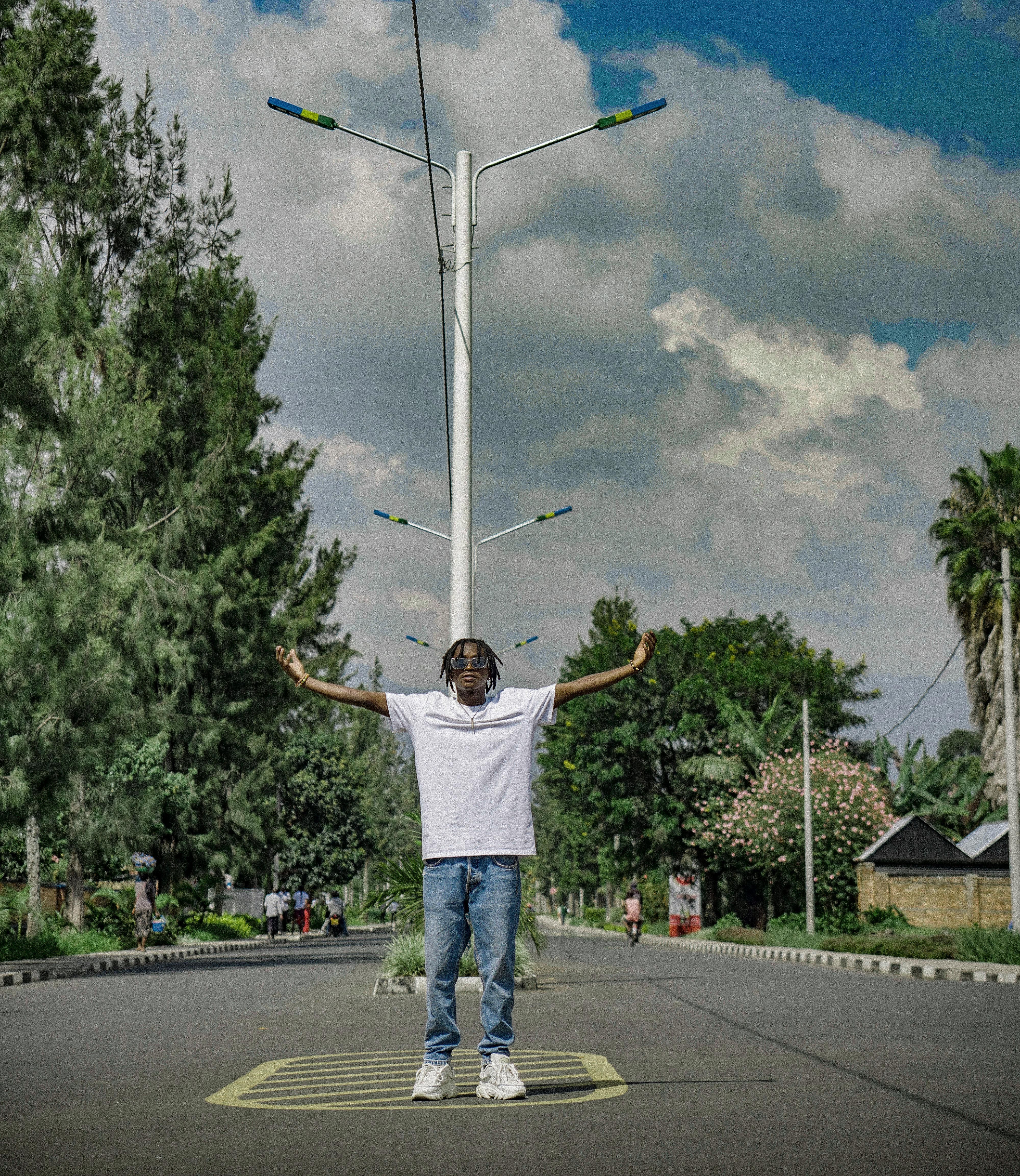 A young man stands on a street with arms raised under a streetlamp, surrounded by lush greenery and an open sky.