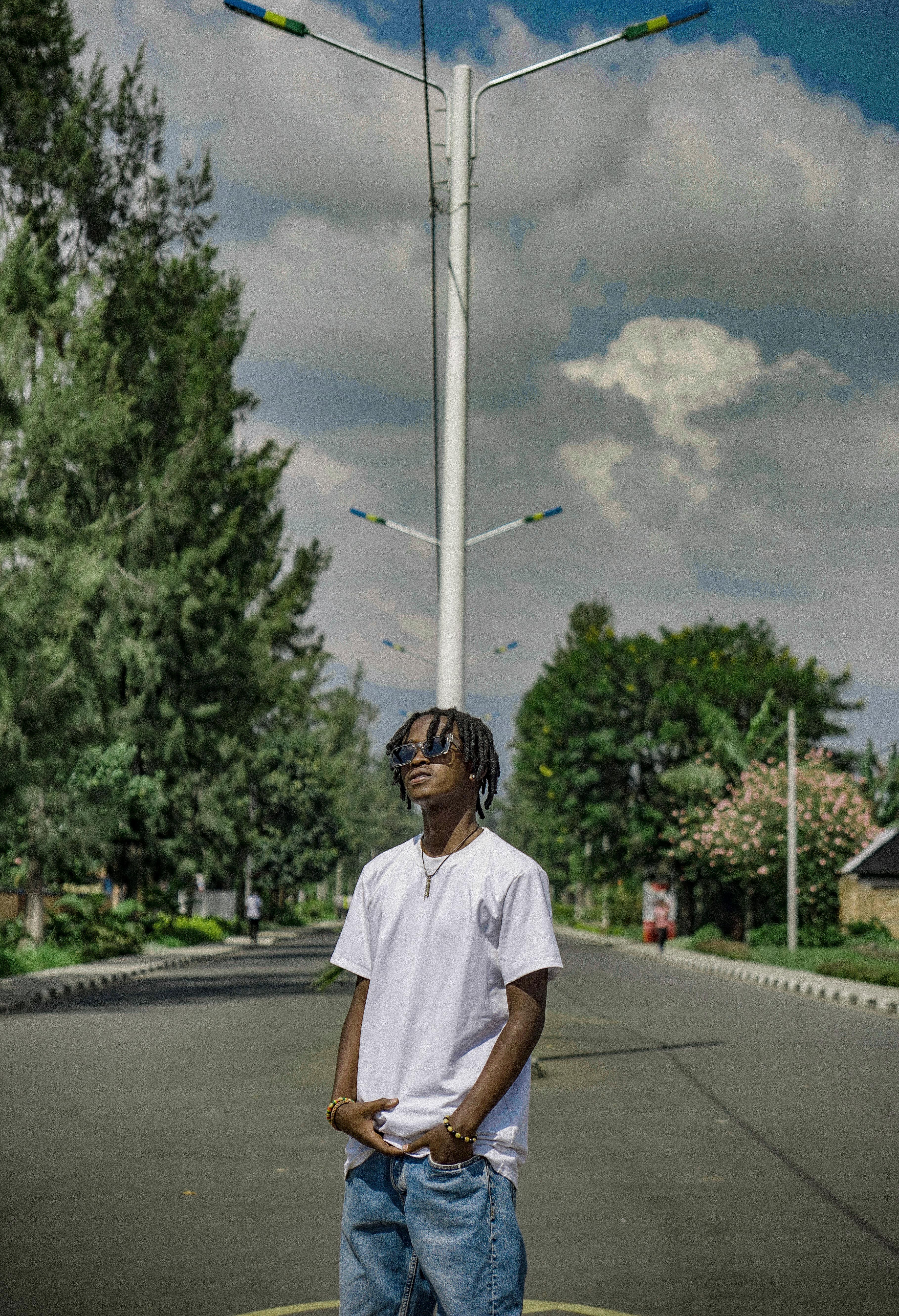 A young adult stands confidently on an empty street surrounded by lush greenery under a bright sky.