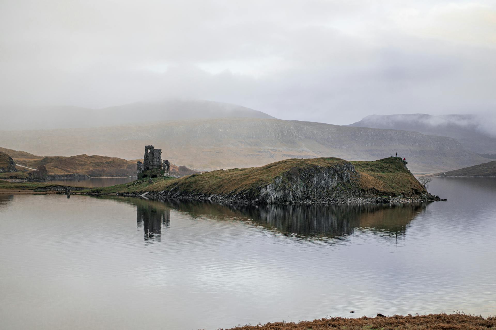 Atmospheric view of a misty Scottish loch with ancient ruin and hillside reflections.