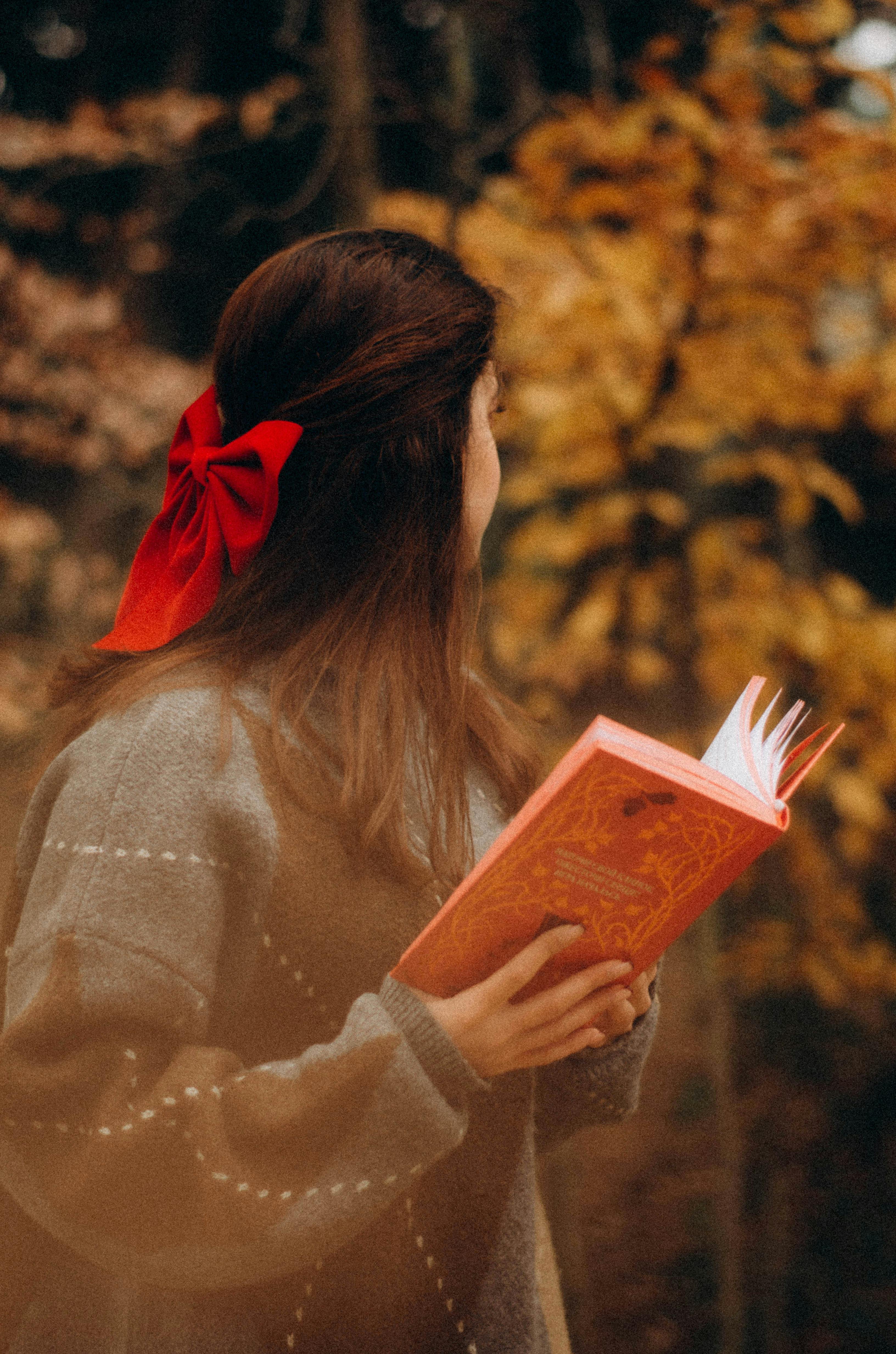 A woman holding a book in an autumn forest, wearing a red bow, scene exudes warm fall vibes.