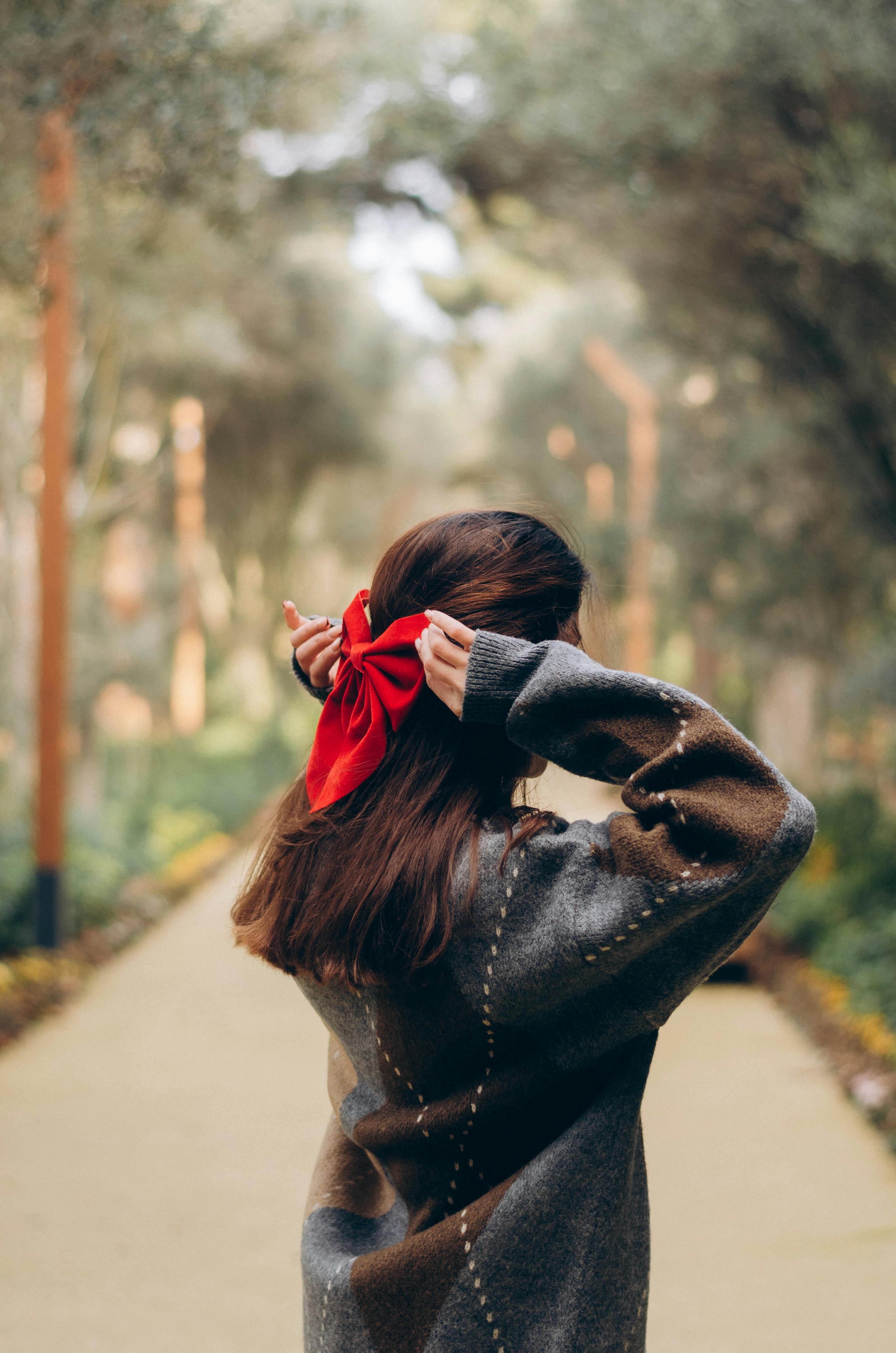 A woman in an oversized sweater adjusts her hair with a red ribbon on a serene park pathway.