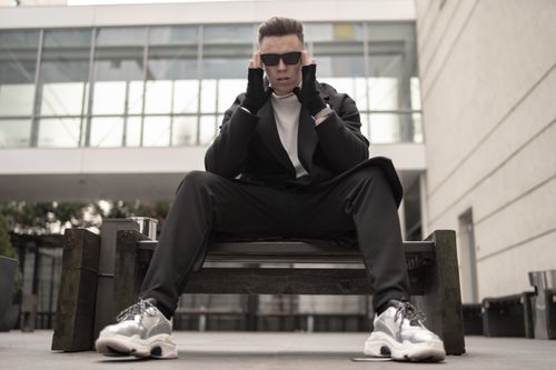 Free Young man in black stylish clothing and sunglasses sits confidently in an urban outdoor setting. Stock Photo