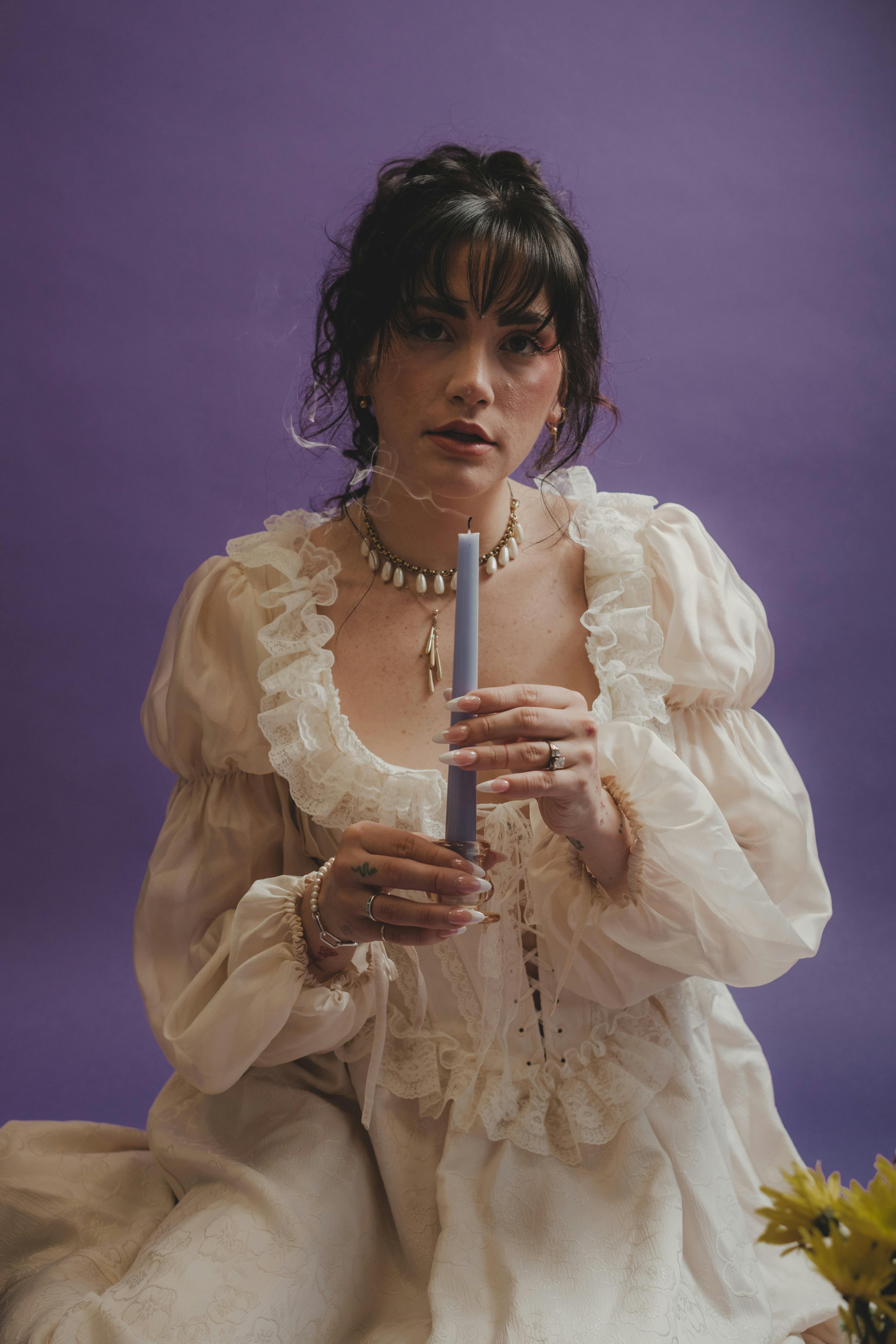 A woman in vintage attire holds a candle against a purple background