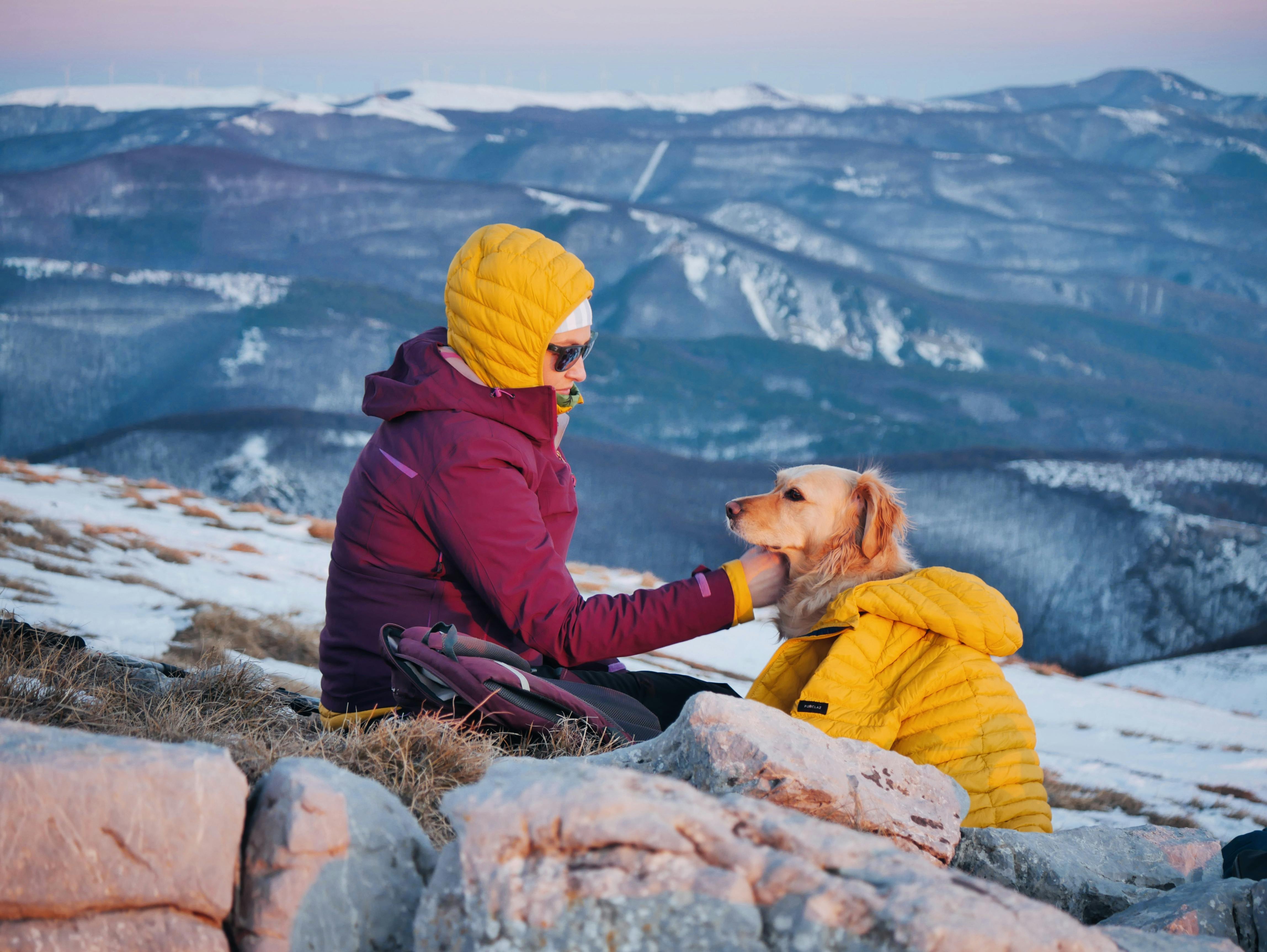 A woman in a warm jacket sits with her dog overlooking a snowy mountain scenery at sunset.