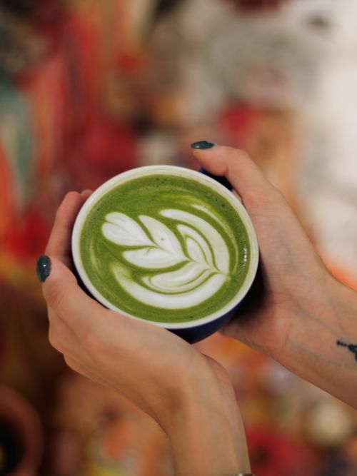 Free Close-up of hands holding a matcha latte with intricate latte art design in a cozy café setting. Stock Photo