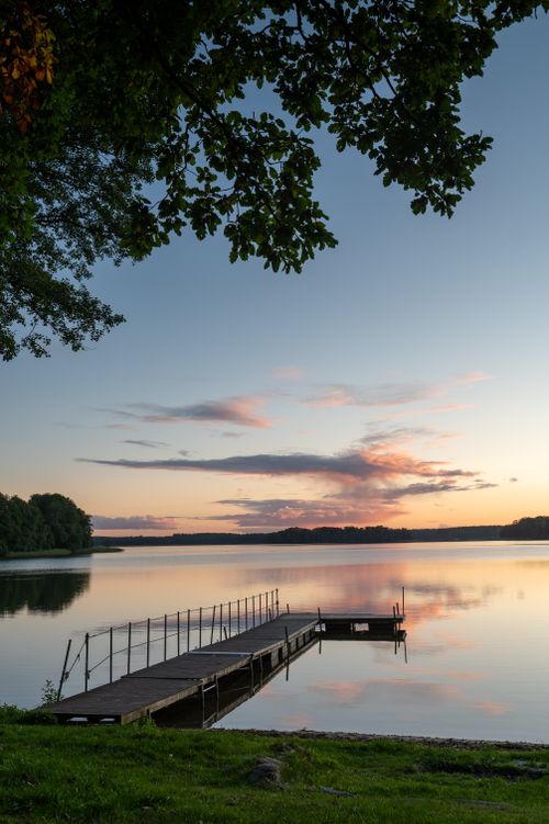 Free Serene summer sunset view over a peaceful lake with a wooden pier and vibrant sky reflections. Stock Photo