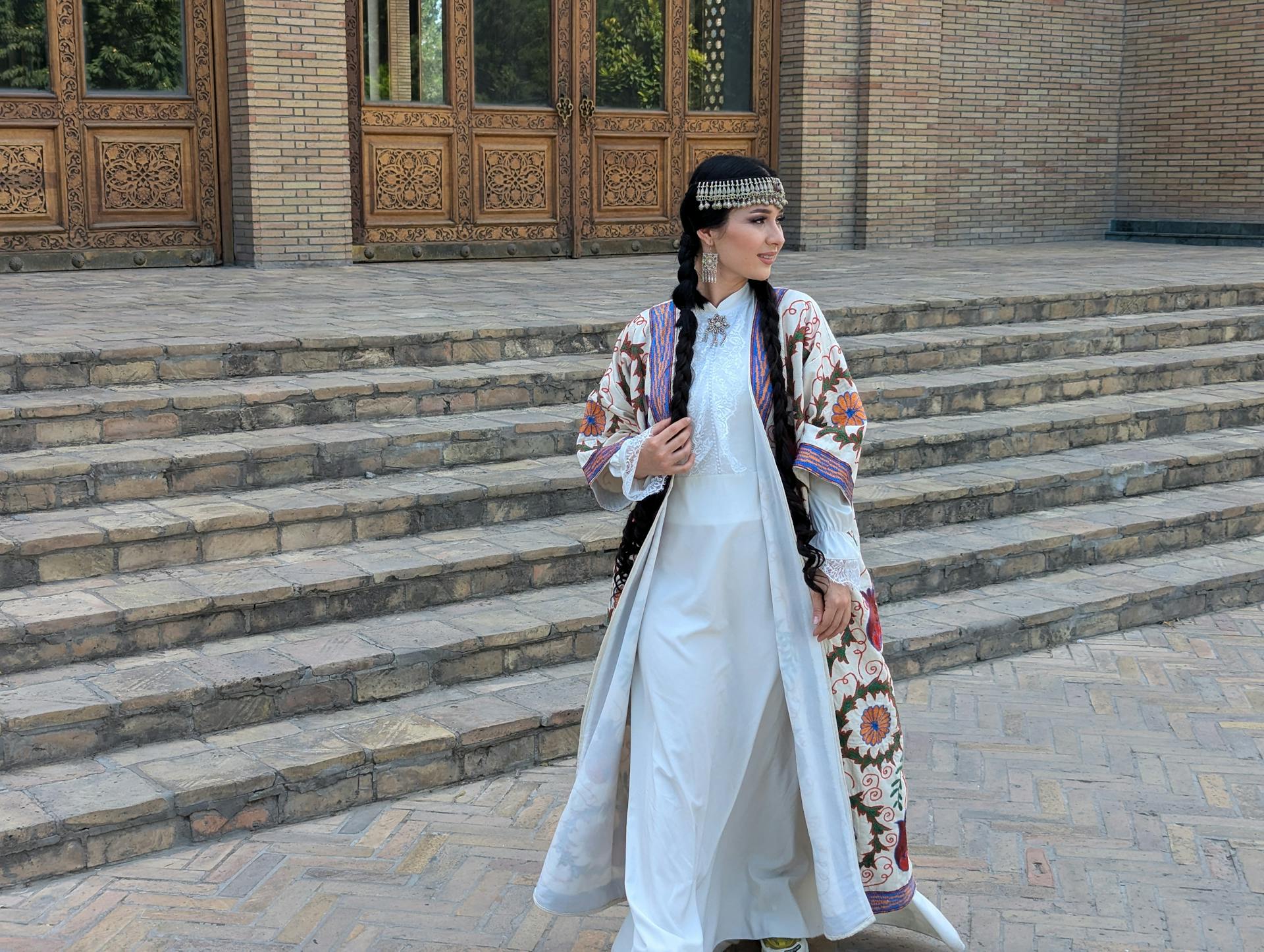 A woman in traditional Uzbek dress poses gracefully outside a historic building with detailed wooden doors.