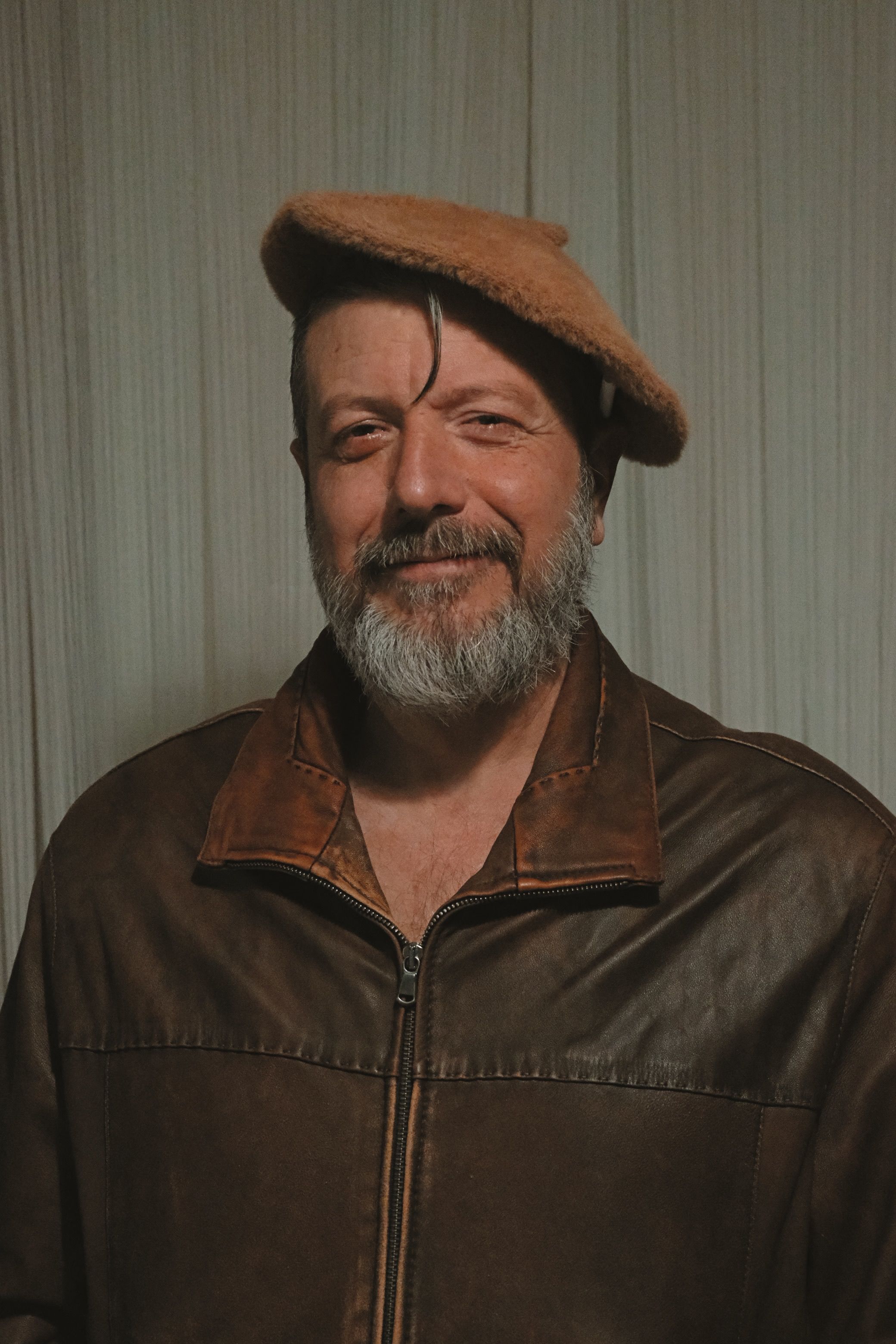A mature man with a beard in a leather jacket and beret, smiling confidently.