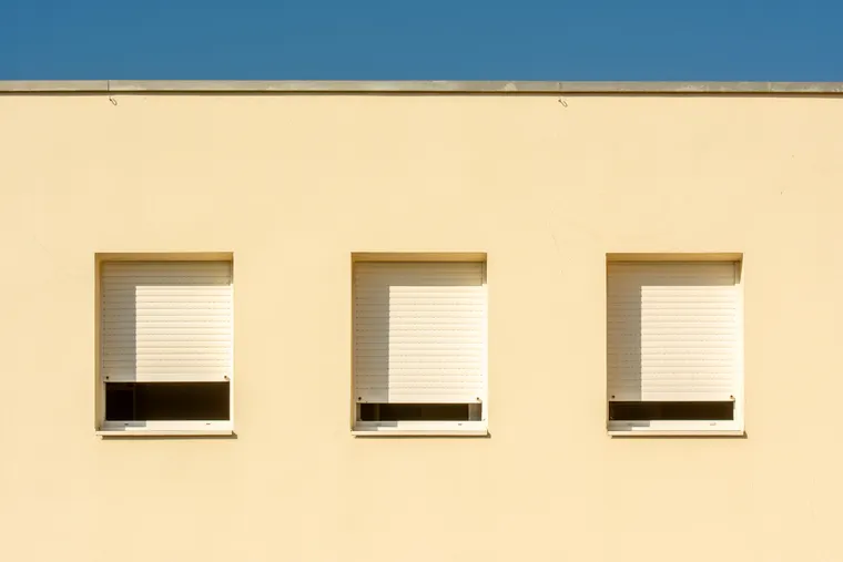 Trois fenêtres d’une façade beige équipées de volets roulants blancs descendus aux trois quarts