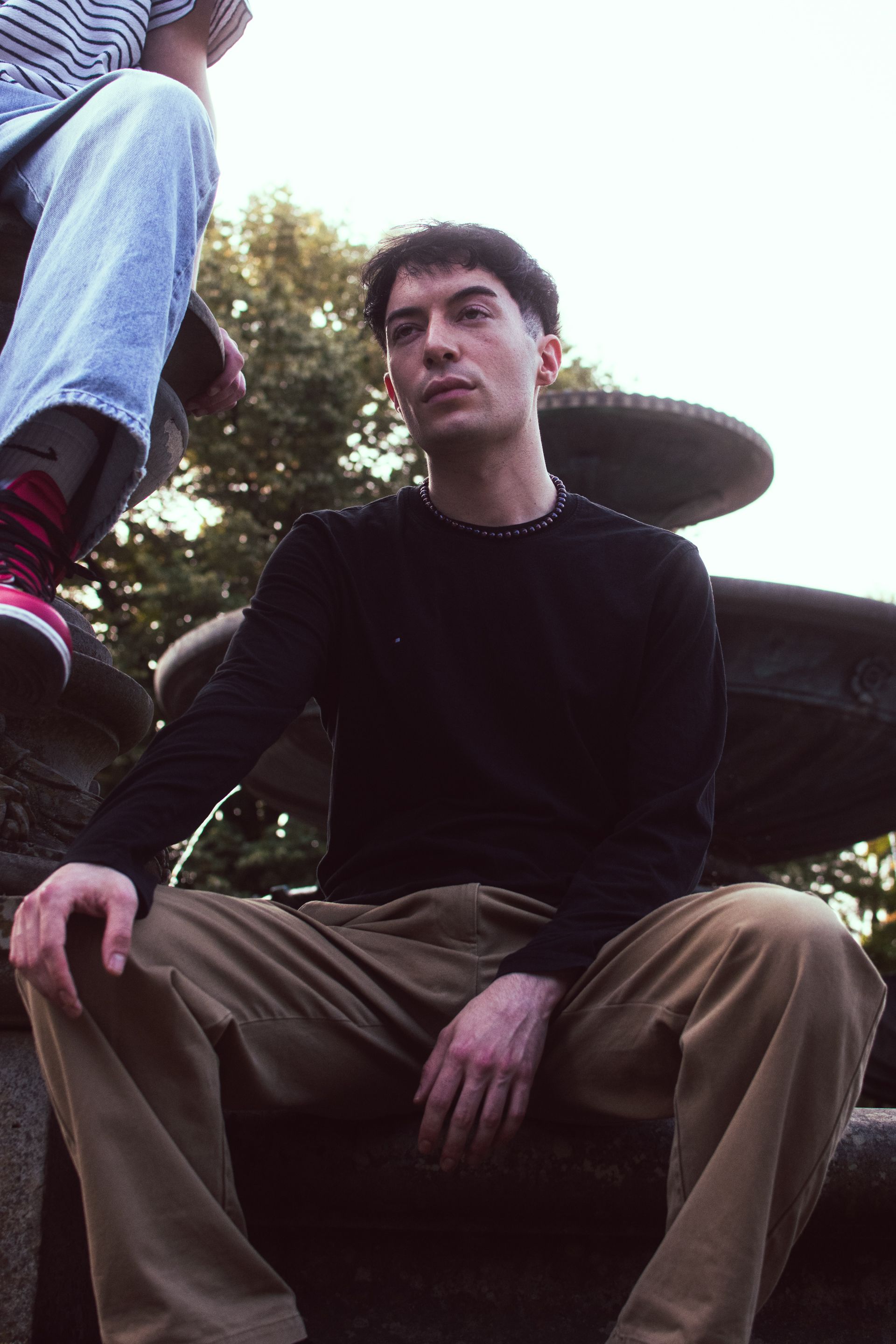 Stylish portrait of a male model sitting by an outdoor fountain with an edgy, dramatic tone.