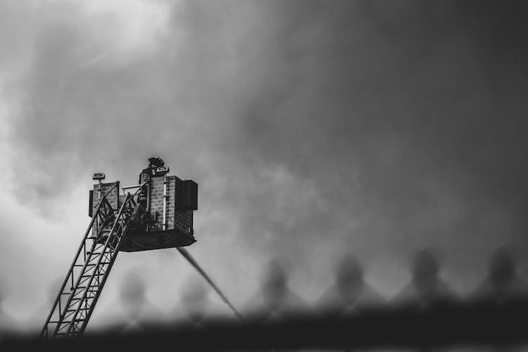 Grayscale Photography Of A Fireman On The Fire Truck Ladder Hosing Water Surrounded By Clouds Of Smoke