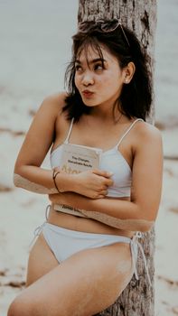 Young woman in white bikini leaning against a tree on a beach, holding a book.