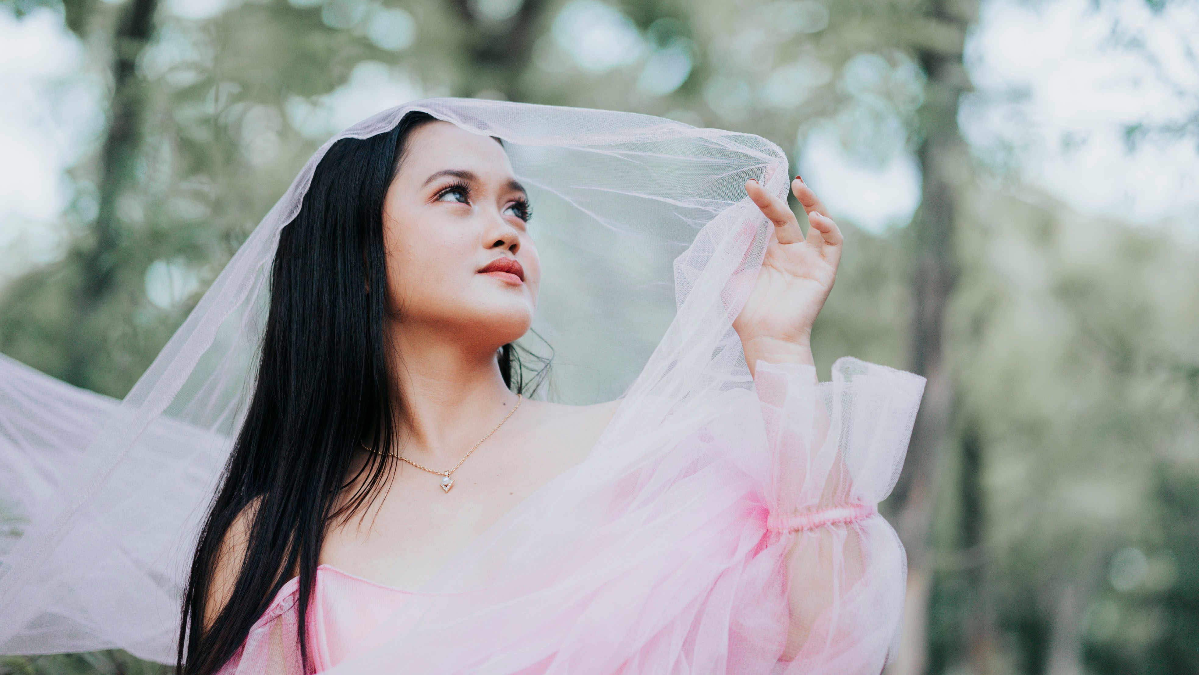 Elegant woman in a pink dress with a veil in a serene outdoor setting.