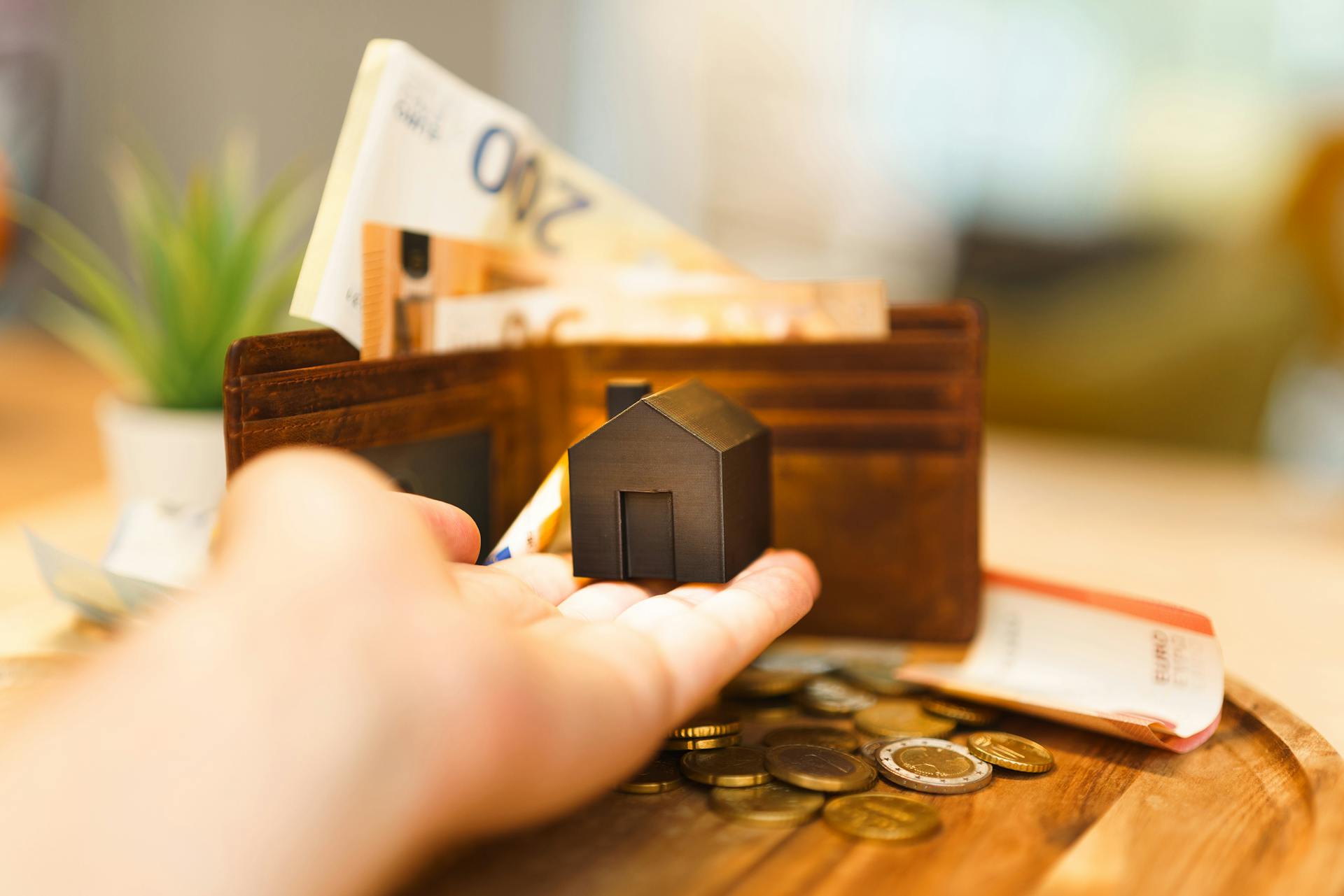 A hand holding a small house model with euro notes and coins nearby, illustrating real estate investment and finance.