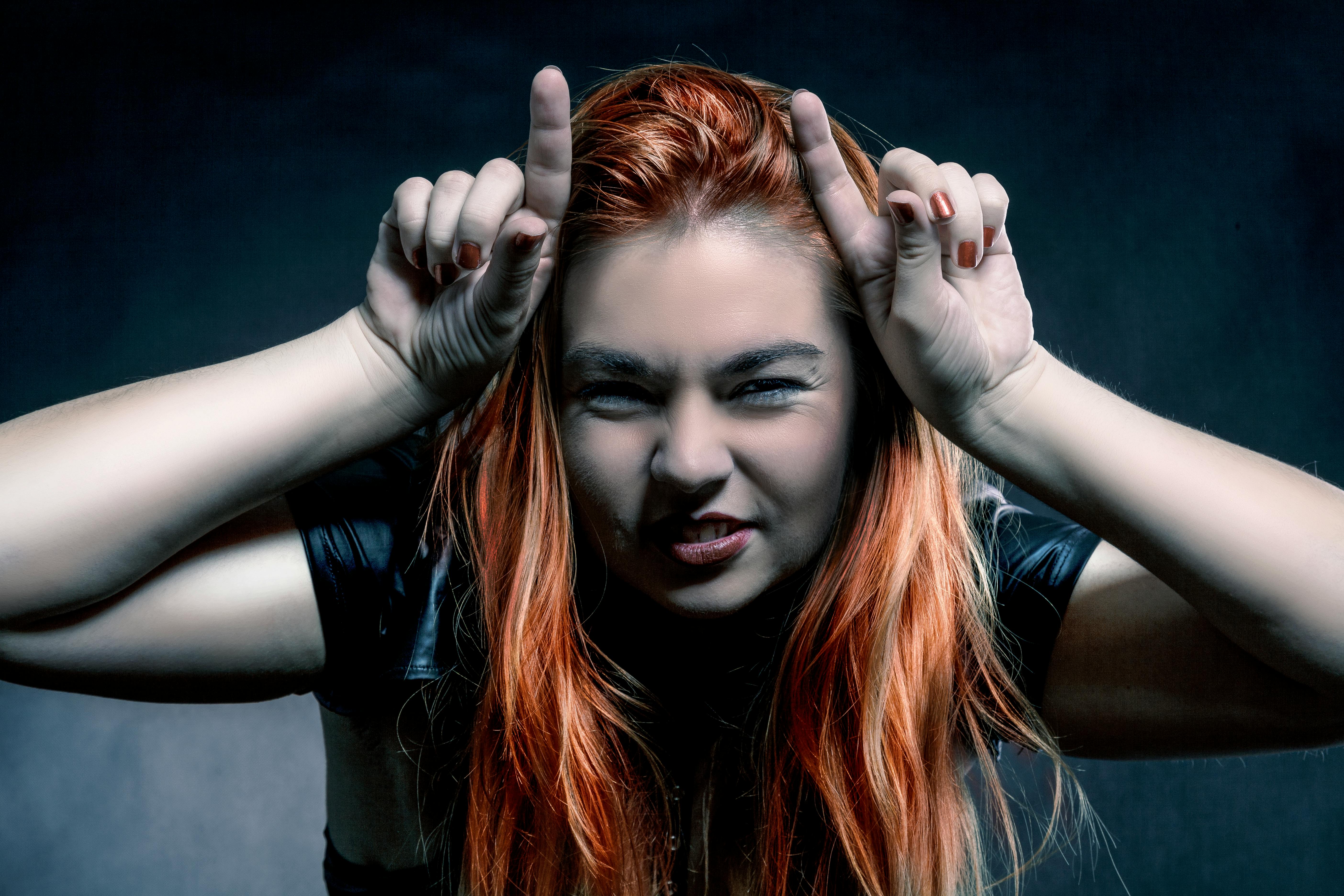 A playful redhead woman with a mischievous expression making a horns gesture against a dark background.