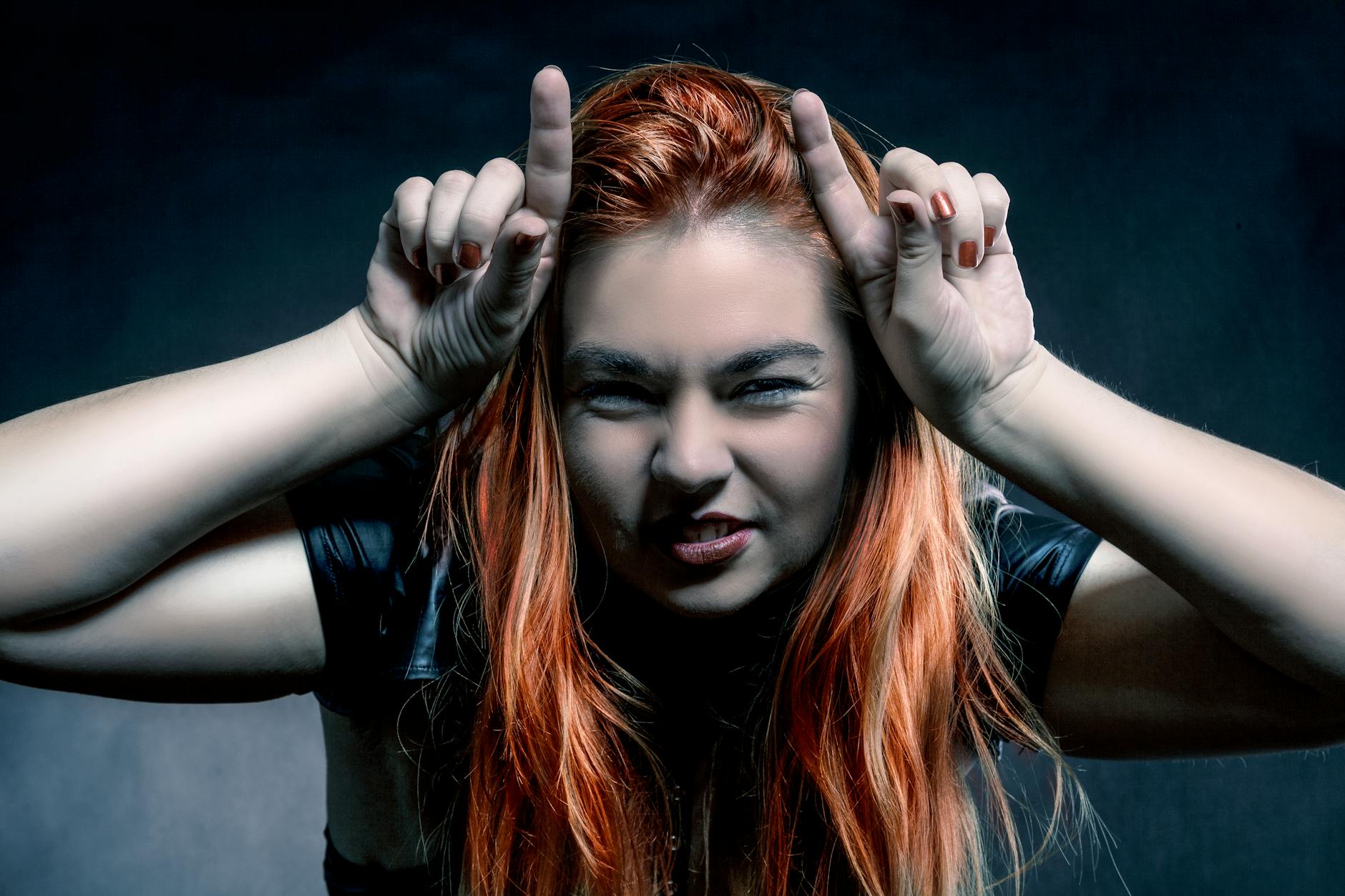 A playful redhead woman with a mischievous expression making a horns gesture against a dark background.