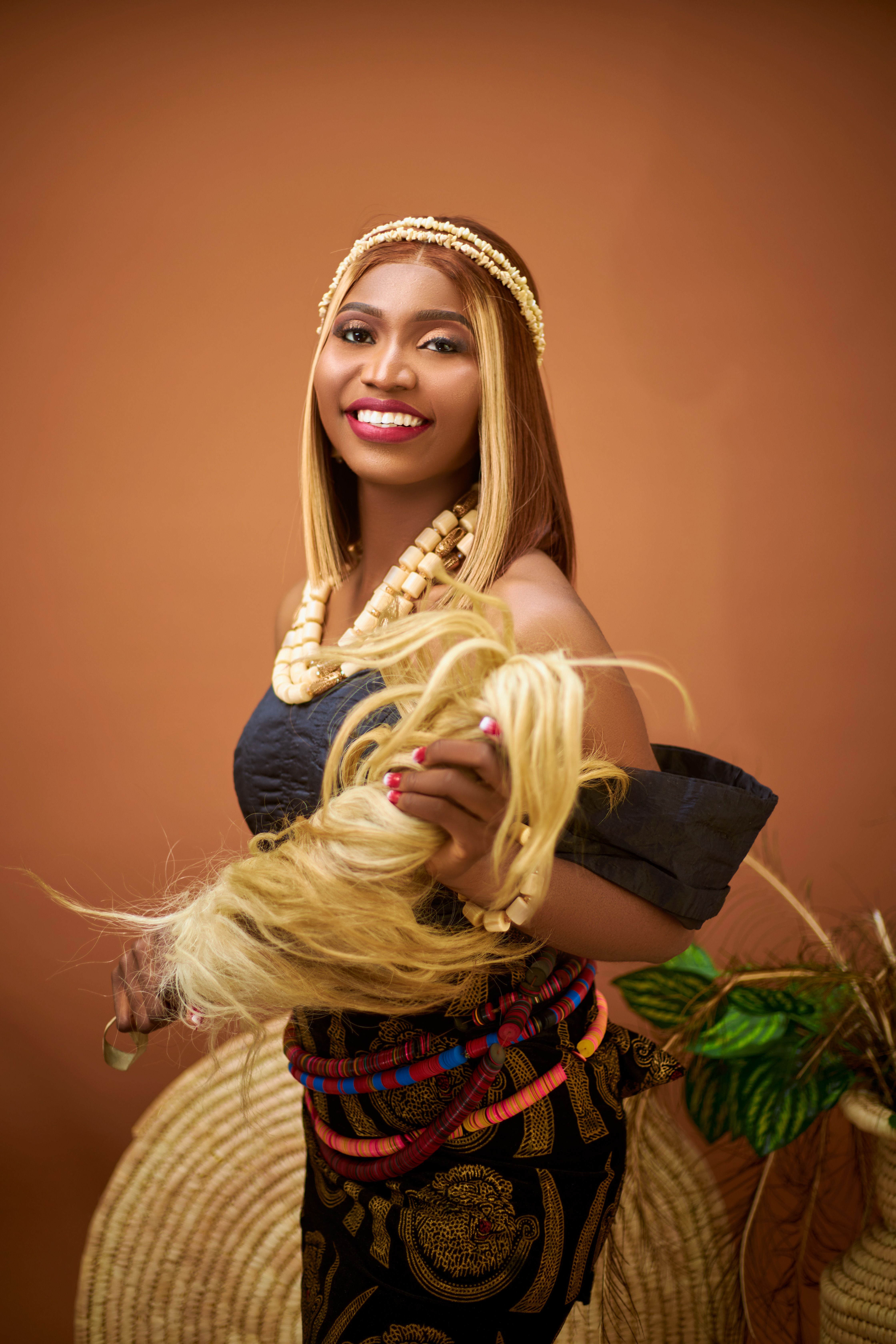 A joyful woman in vibrant traditional clothing holding a blonde hair accessory against a warm-toned background.