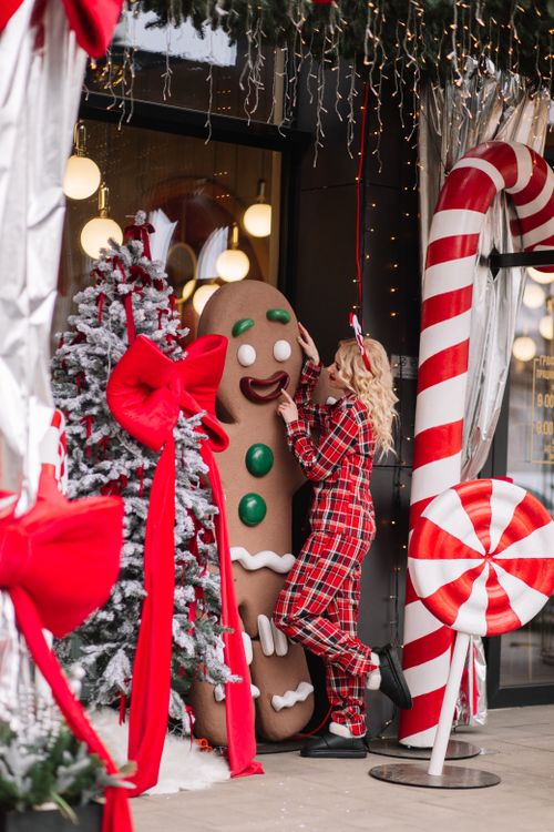 Free A woman in plaid pajamas interacting with a large gingerbread man decoration amidst Christmas decor. Stock Photo