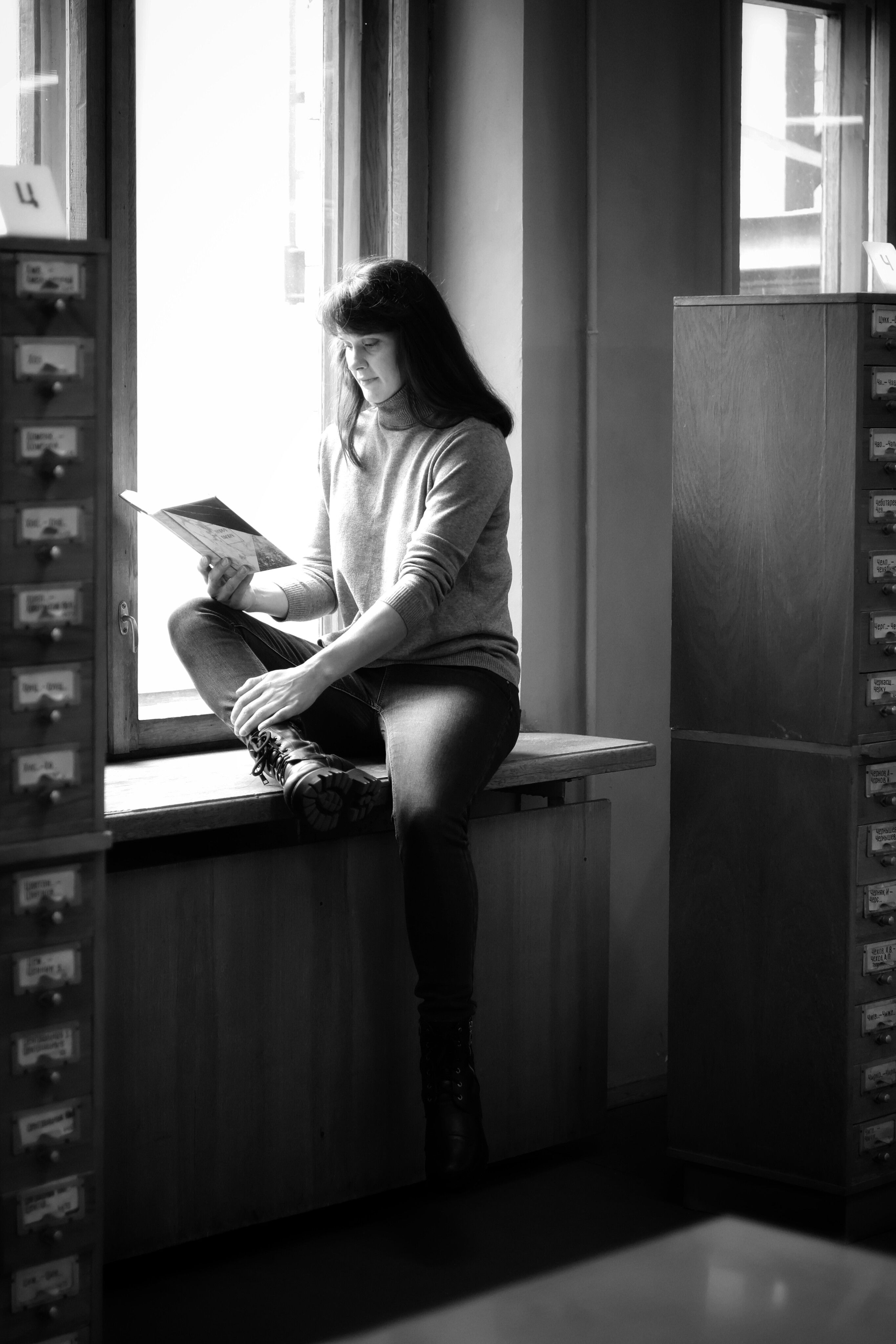 A woman sits by a library window reading a book in a calm, black and white setting.