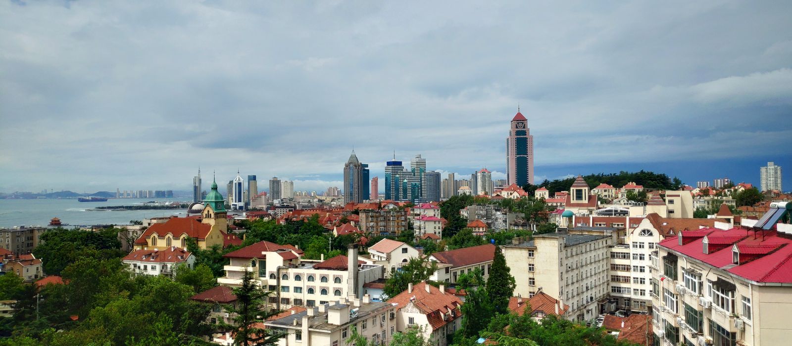 A scenic view of Qingdao's skyline with historical buildings and modern skyscrapers under a cloudy sky. — flights from Shanghai to Qingdao