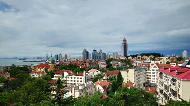 A scenic view of Qingdao's skyline with historical buildings and modern skyscrapers under a cloudy sky. — lounges at Qingdao Airport