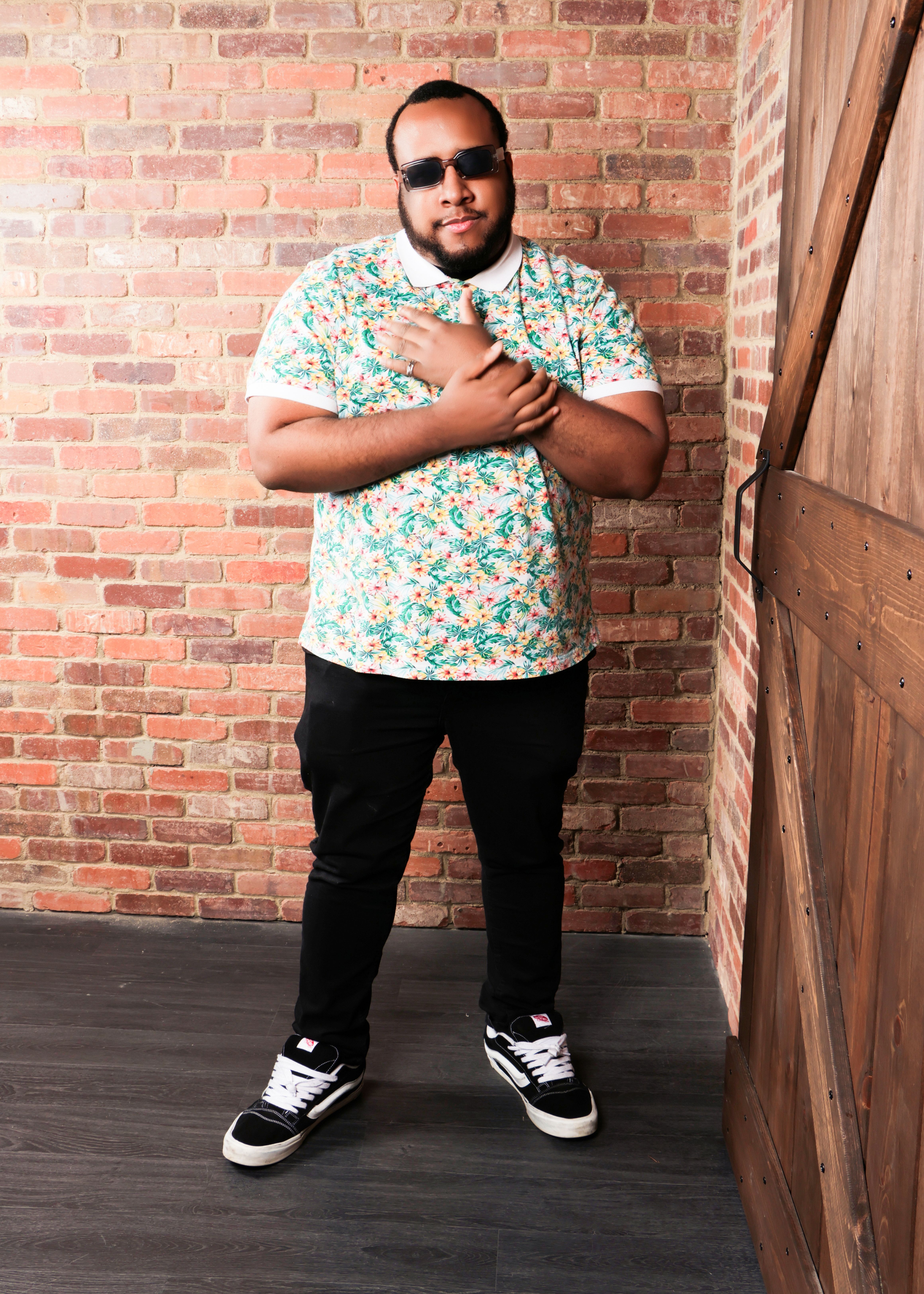 Man with sunglasses in floral shirt posing indoors against a brick wall