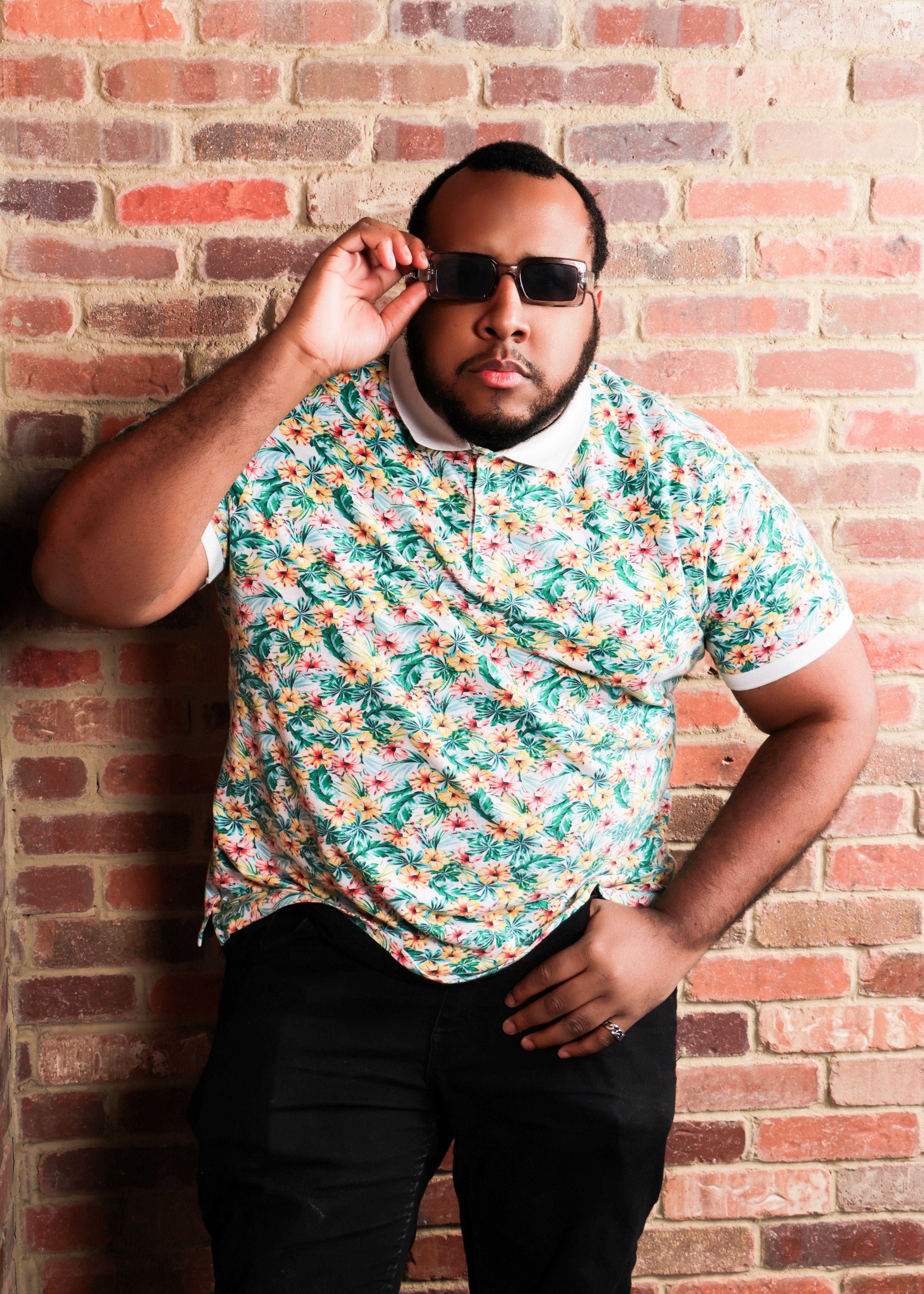 A fashionable man poses confidently against a brick wall wearing sunglasses and a floral shirt.