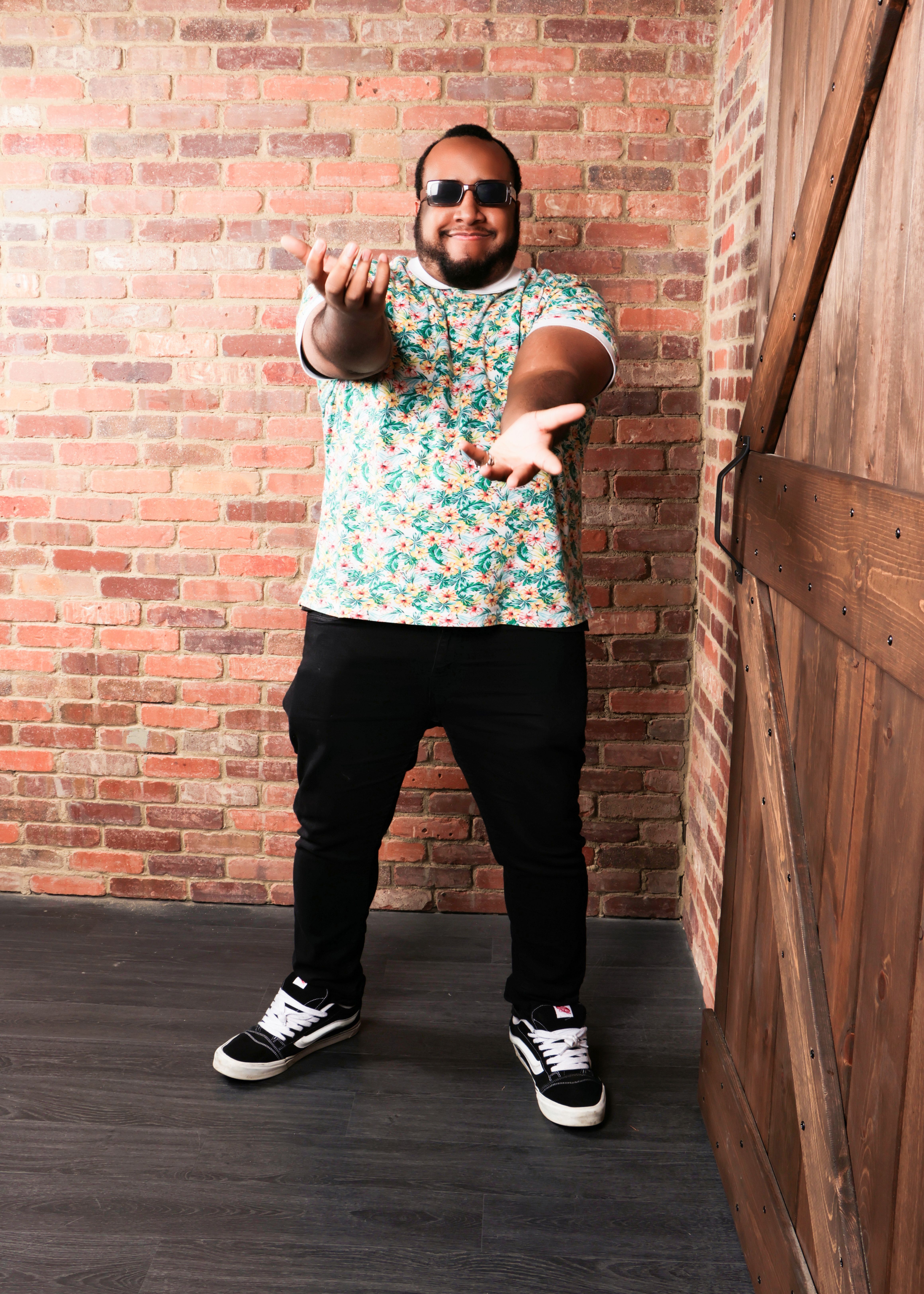 A fashionable man poses with a smile in front of a rustic brick wall, exuding confidence.