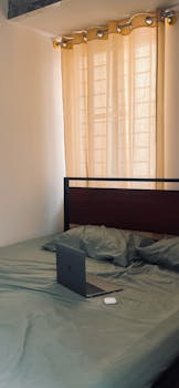 A cozy home office setup with laptop on bed, curtain filtered sunlight streaming in, creating a calm workspace.