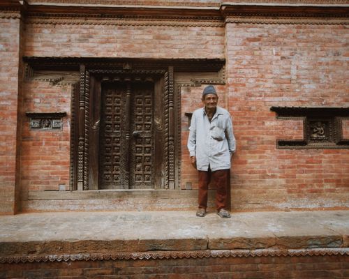 Free Elderly man poses by a traditional brick building with ornate wooden door. Stock Photo