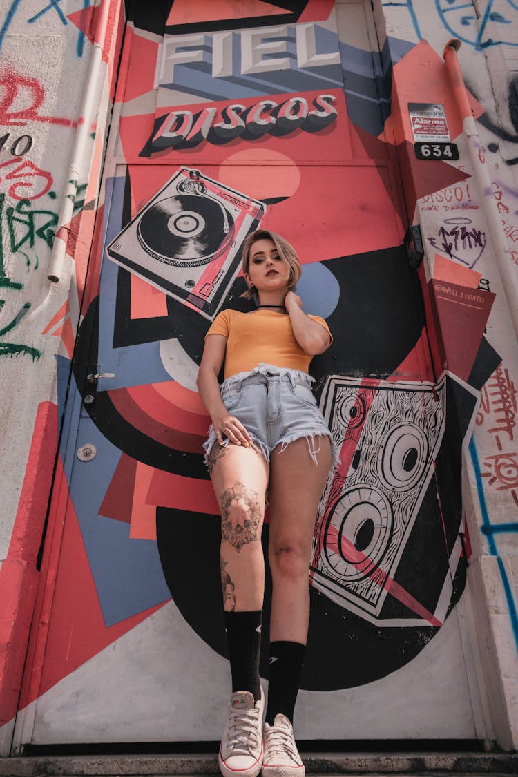 Low Angle Photo Of Tattooed Woman In Orange T-shirt And Blue Denim Shorts Leaning On Graffiti Door