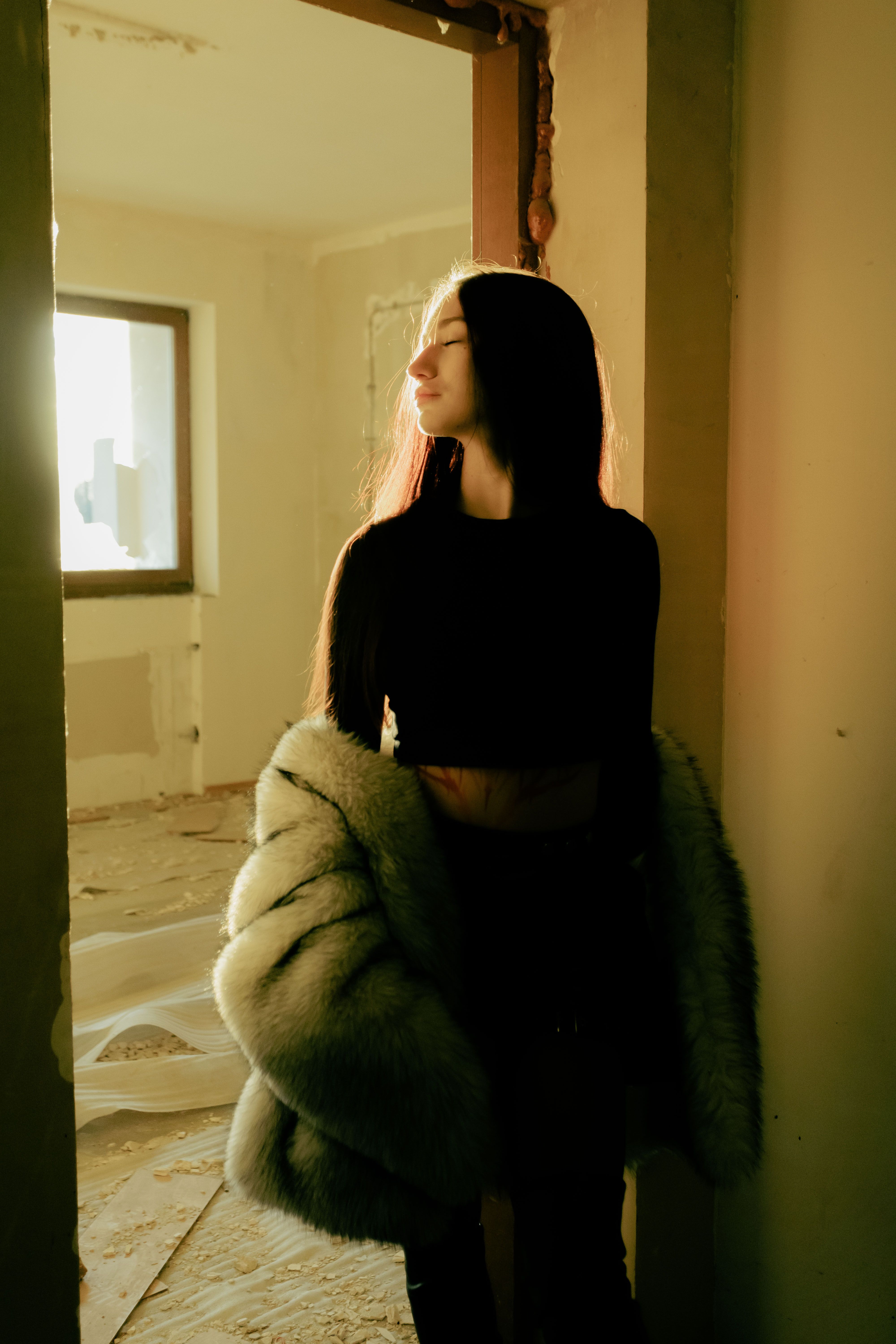 A young woman in a fur jacket stands in a sunlit room, embracing warmth.