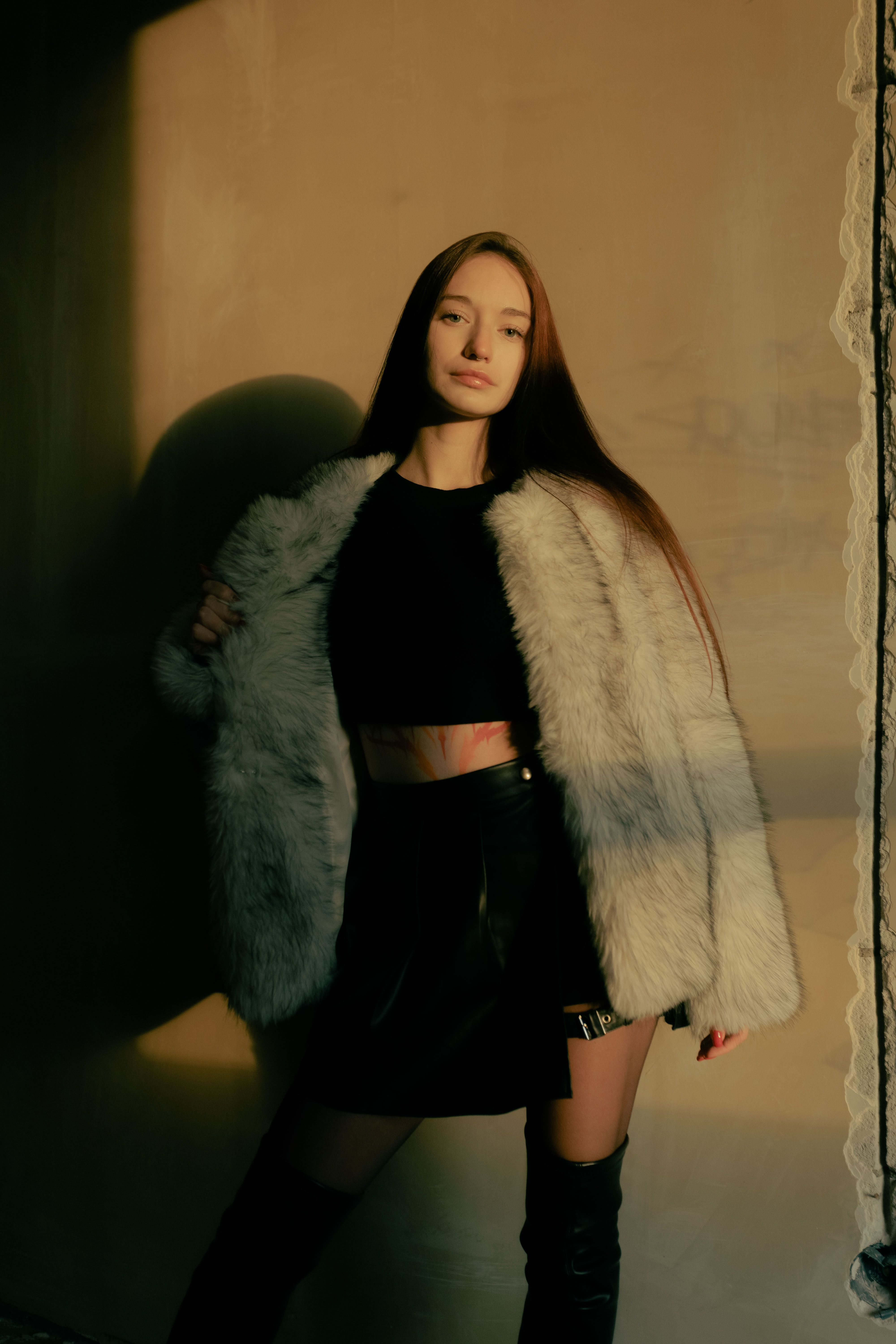 Confident woman posing in fur coat with warm sunlight on a beige wall.