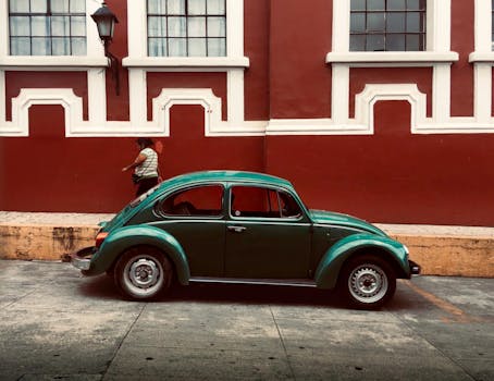 Classic green Beetle car parked by a red brick wall with a pedestrian in motion.