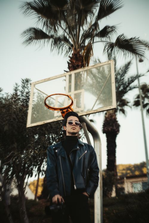 Free Young adult male posing by basketball hoop with palm trees, exuding a fashion-forward vibe. Stock Photo