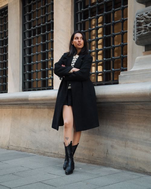 Free A woman in a stylish coat confidently leaning against a decorative urban wall. Stock Photo