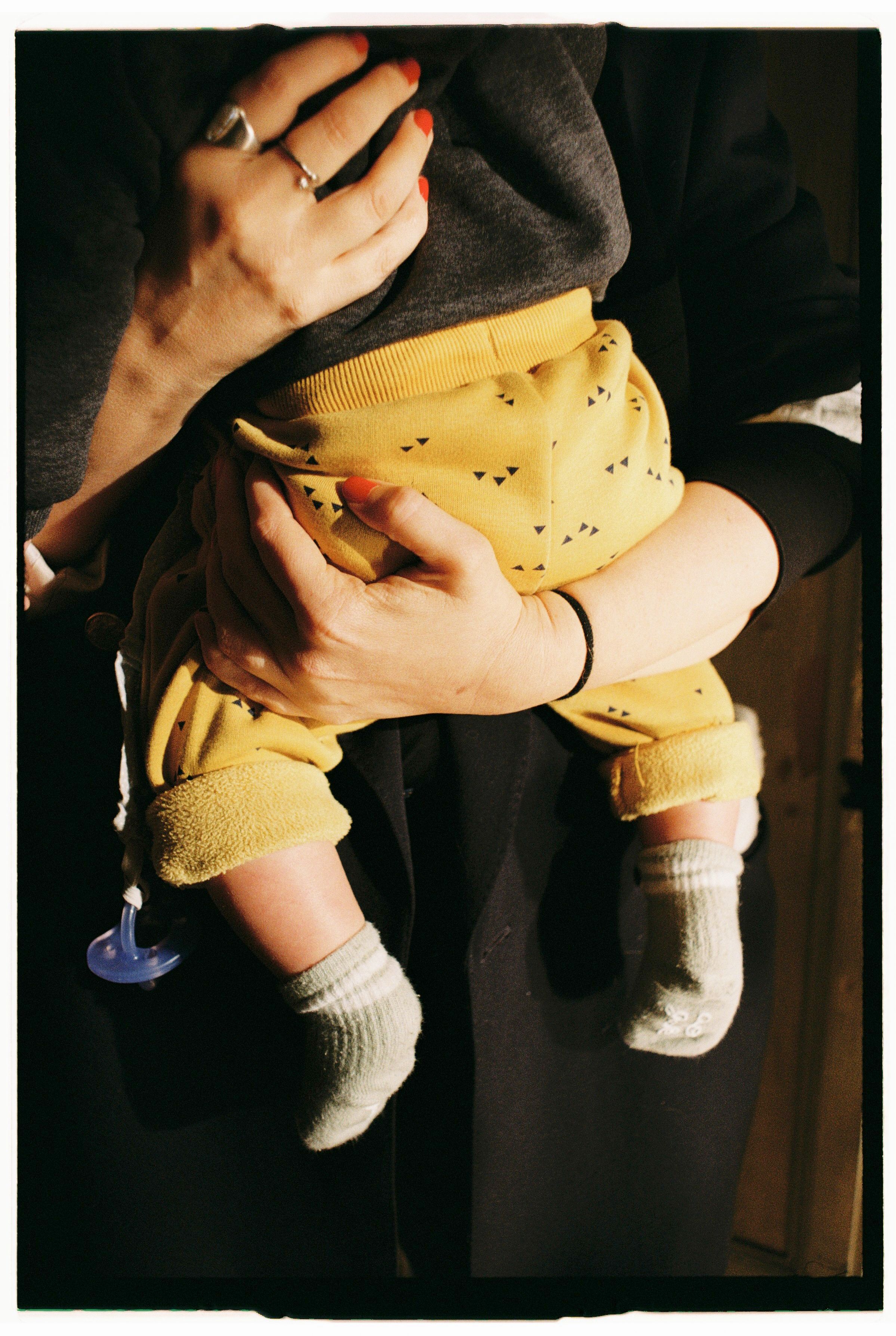 A mother lovingly holds her baby with yellow pants indoors with warm lighting.