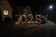 Sparkler light painting forming '2025' in a festive night street scene.