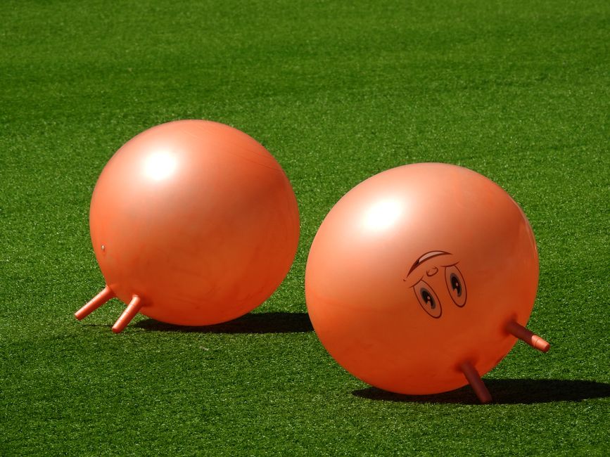 Two orange hopper balls with faces resting on a vibrant green turf, under bright daylight.